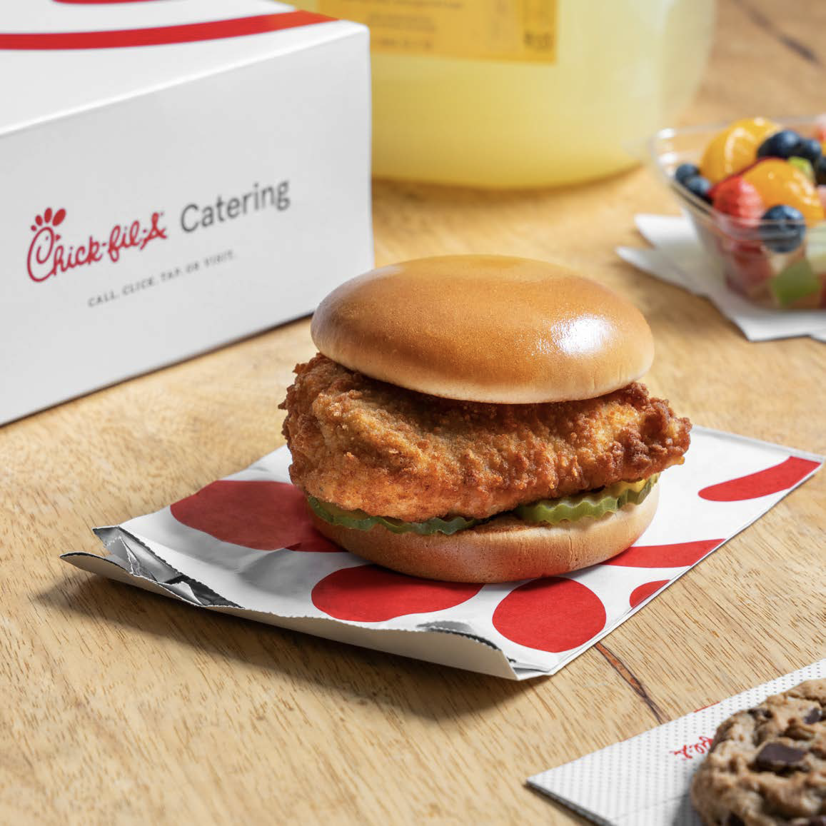 A Chick-fil-A chicken sandwich with pickles in a soft bun, resting on a red and white paper wrapper on a wooden table, with a Chick-fil-A catering box and a fruit cup in the background.