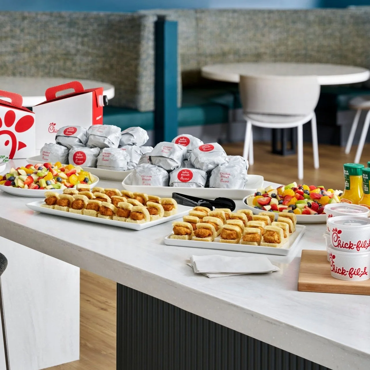 A buffet table with Chick-fil-A sandwiches wrapped in paper, bowls of fruit salad, small sandwiches, and cups of drinks in a bright indoor setting.