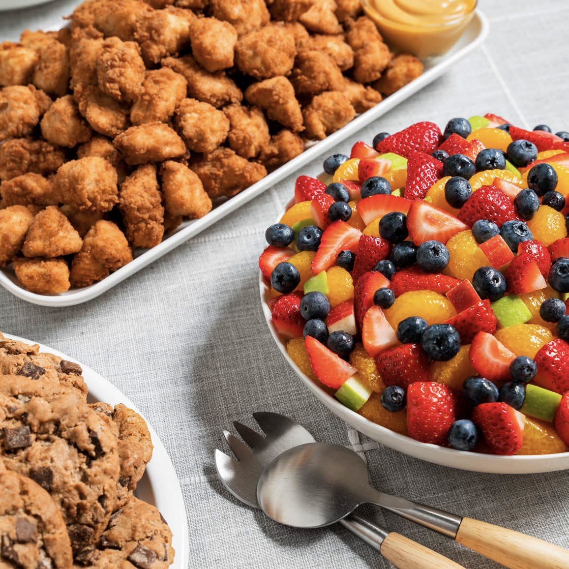 A serving tray of fried chicken bites, a bowl of mixed fresh fruit including strawberries, blueberries, kiwi, and oranges, and chocolate chip cookies with a spoon and fork on a gray tablecloth.