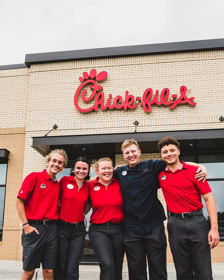 five Chick-fil-A employees in uniform standing outside the restaurant, smiling and posing together.