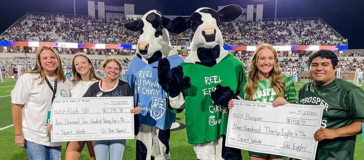 Group of six people on a football field, two dressed as cows, all smiling and holding large checks, with a large crowd in the stands behind them.