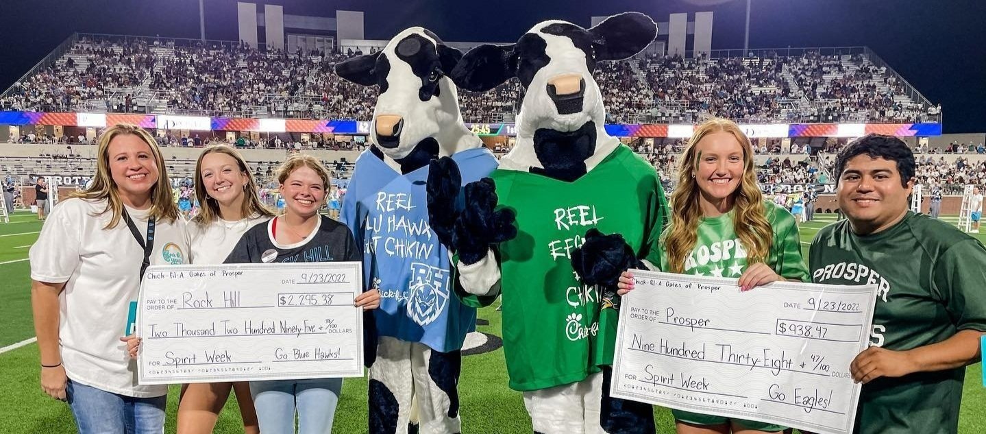 Group of students and cheerleaders at a sports stadium, holding oversized checks for Spirit Week donations, with cow mascots in the center.