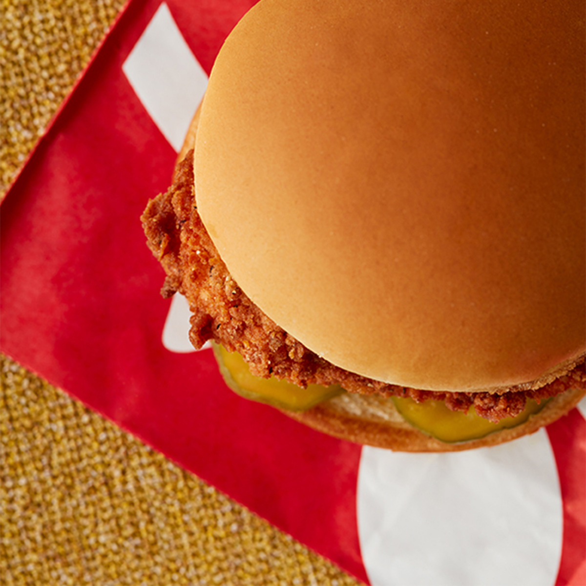 Close-up of a fried chicken sandwich with pickles on a bun, on red and white branded paper, on a textured surface.