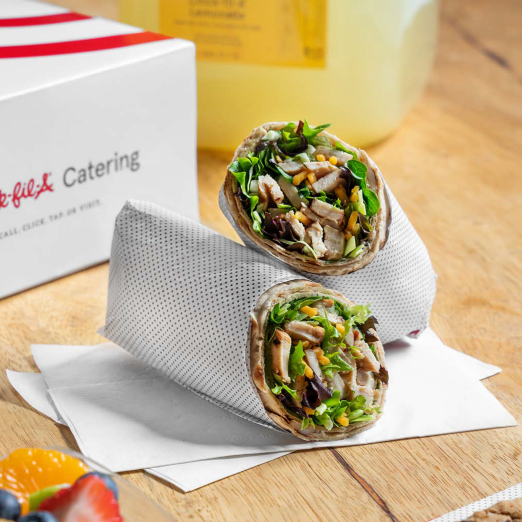Two wraps filled with grilled chicken, lettuce, and shredded cheese, wrapped in white paper, on a wooden surface with a Chick-fil-A catering box and a yellow container in the background.