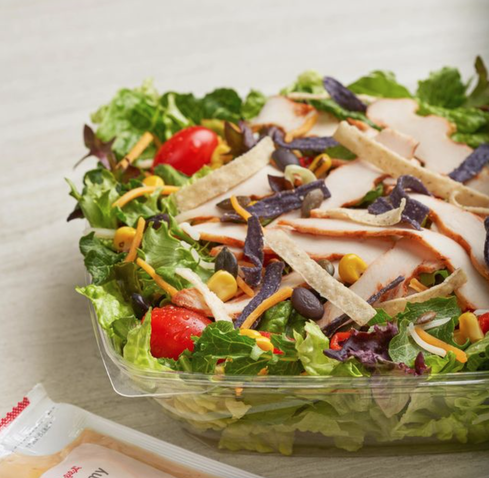 Fresh salad with lettuce, cherry tomatoes, shredded cheese, grilled chicken slices, black beans, corn, and tortilla strips in a clear plastic container.