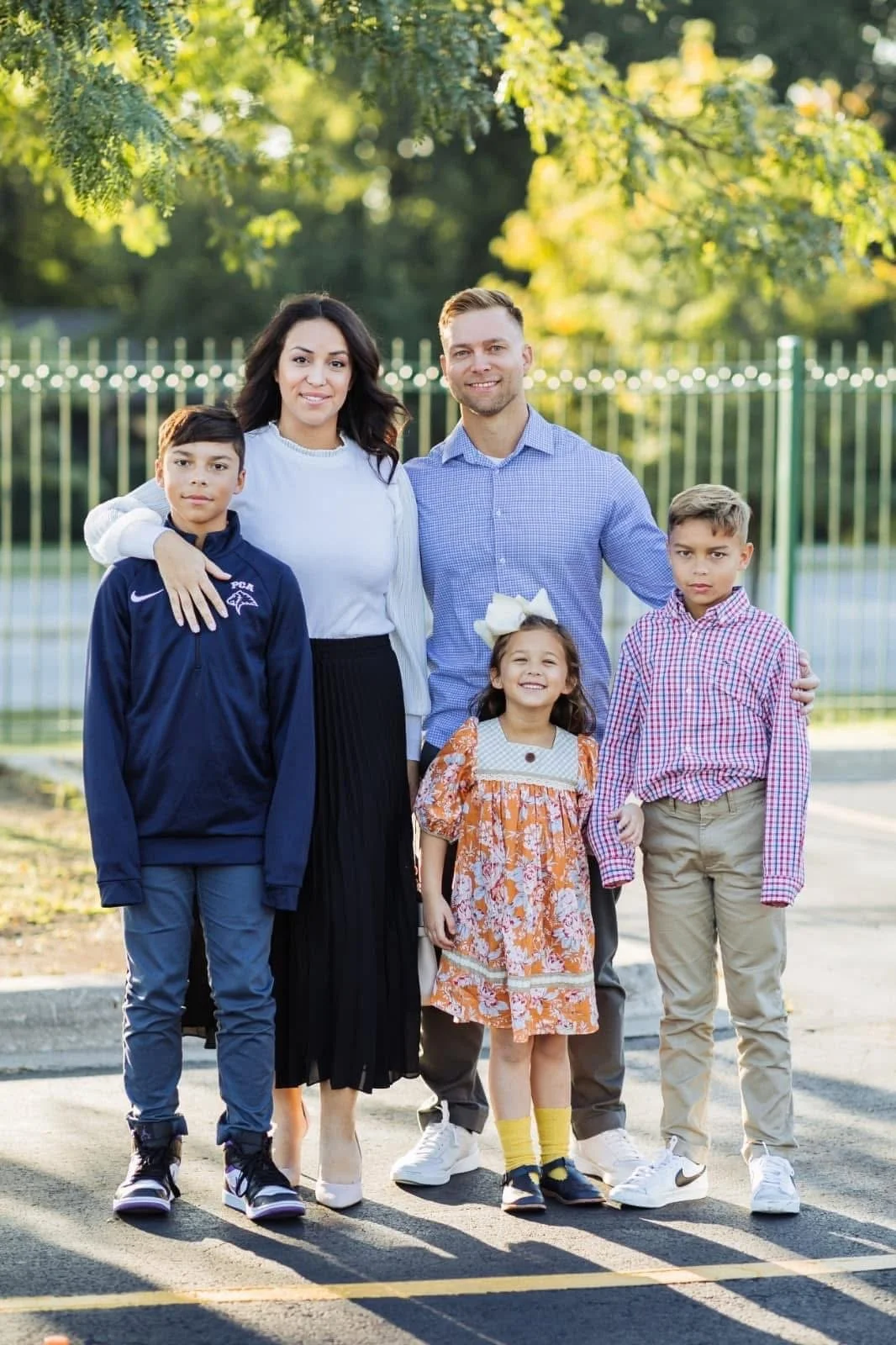 A family of six standing outdoors on a sunny day, children with varied clothing styles, smiling, in front of a green fence with trees.