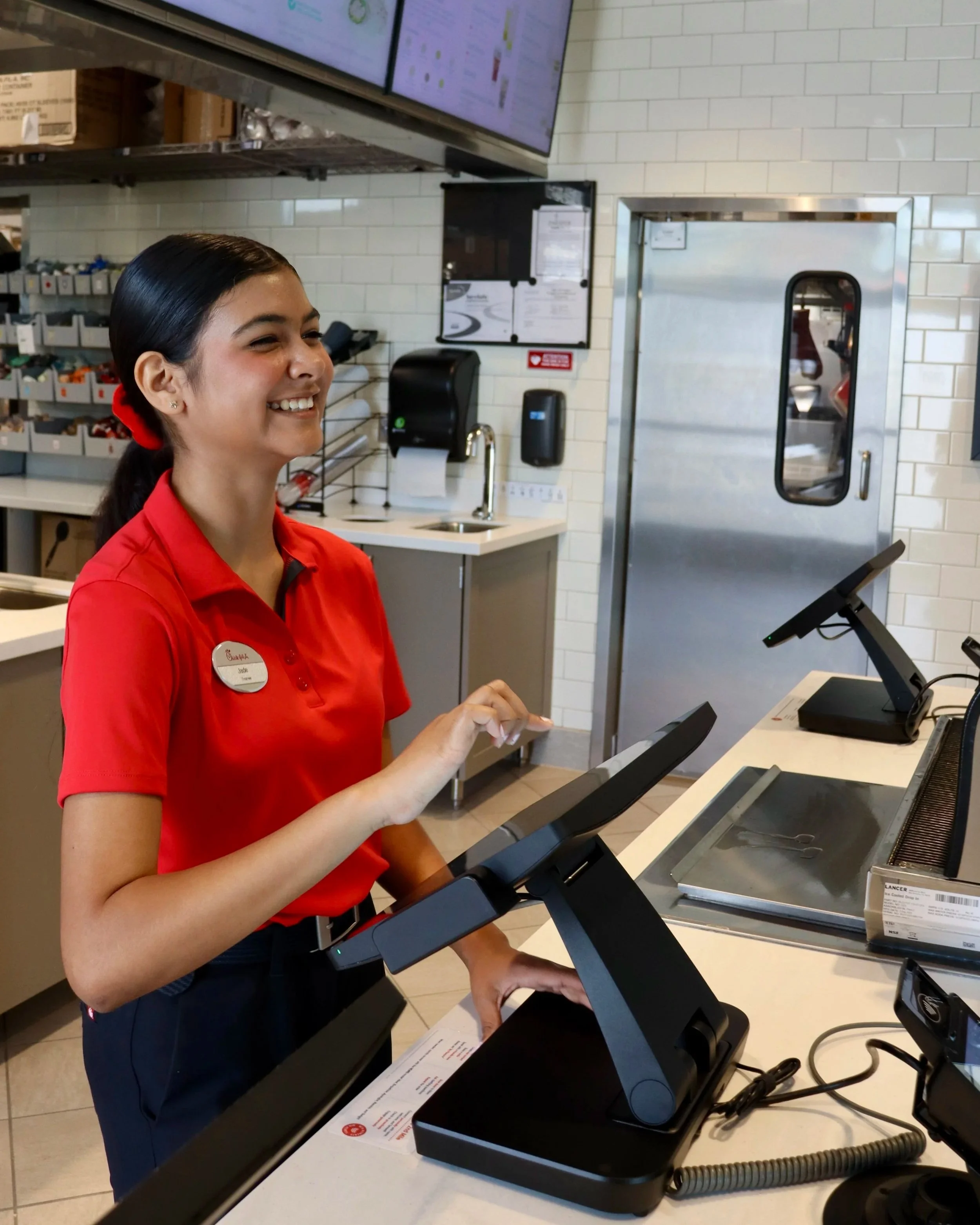 A smiling female employee wearing a red uniform working at a checkout station in a retail store, with a touchscreen register and a barcode scanner on the counter.