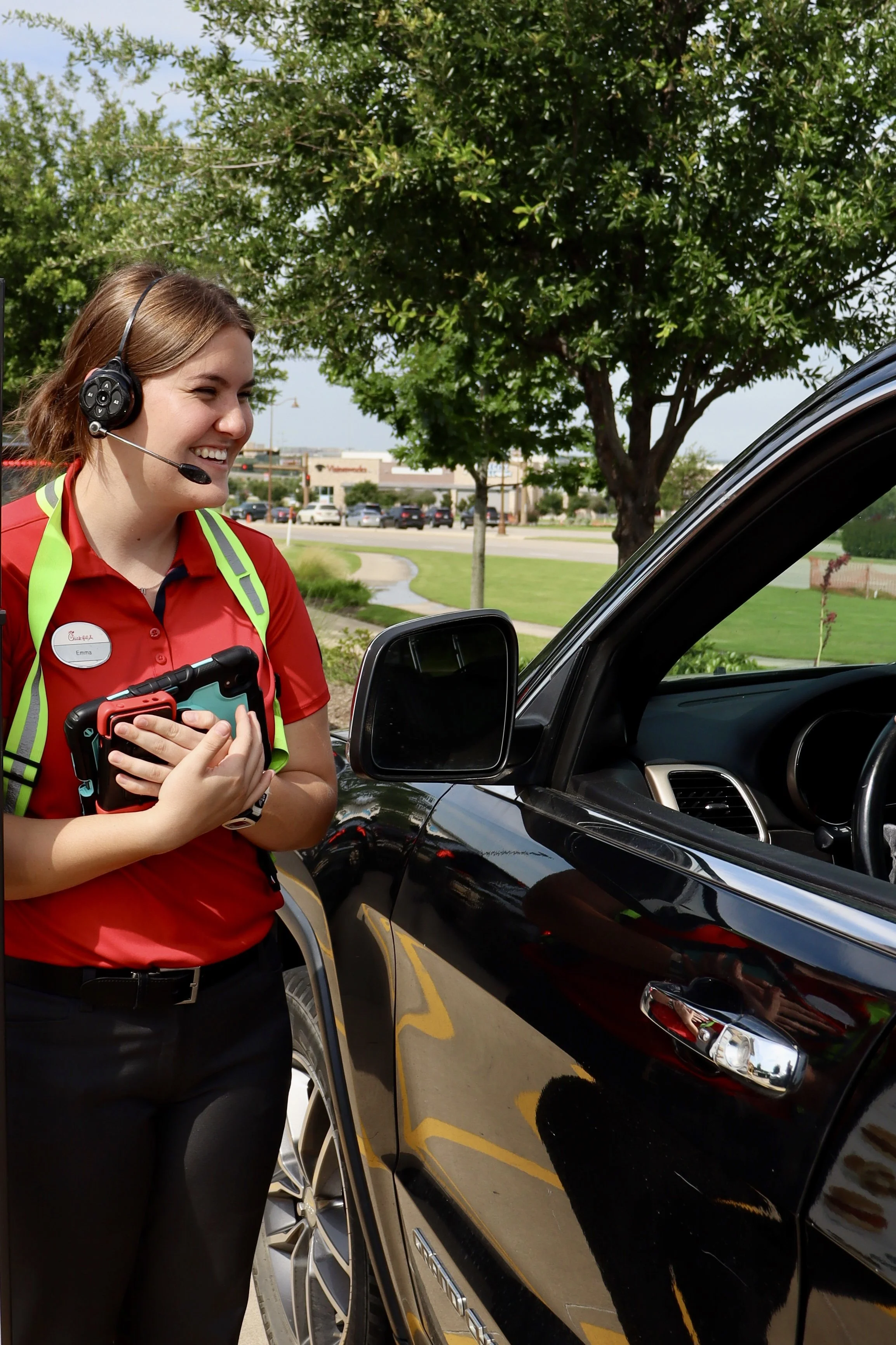 A smiling woman in a red uniform with a name tag that reads 'Emma' and wearing a headset, standing next to a black vehicle in a parking lot with trees and a shopping center in the background.