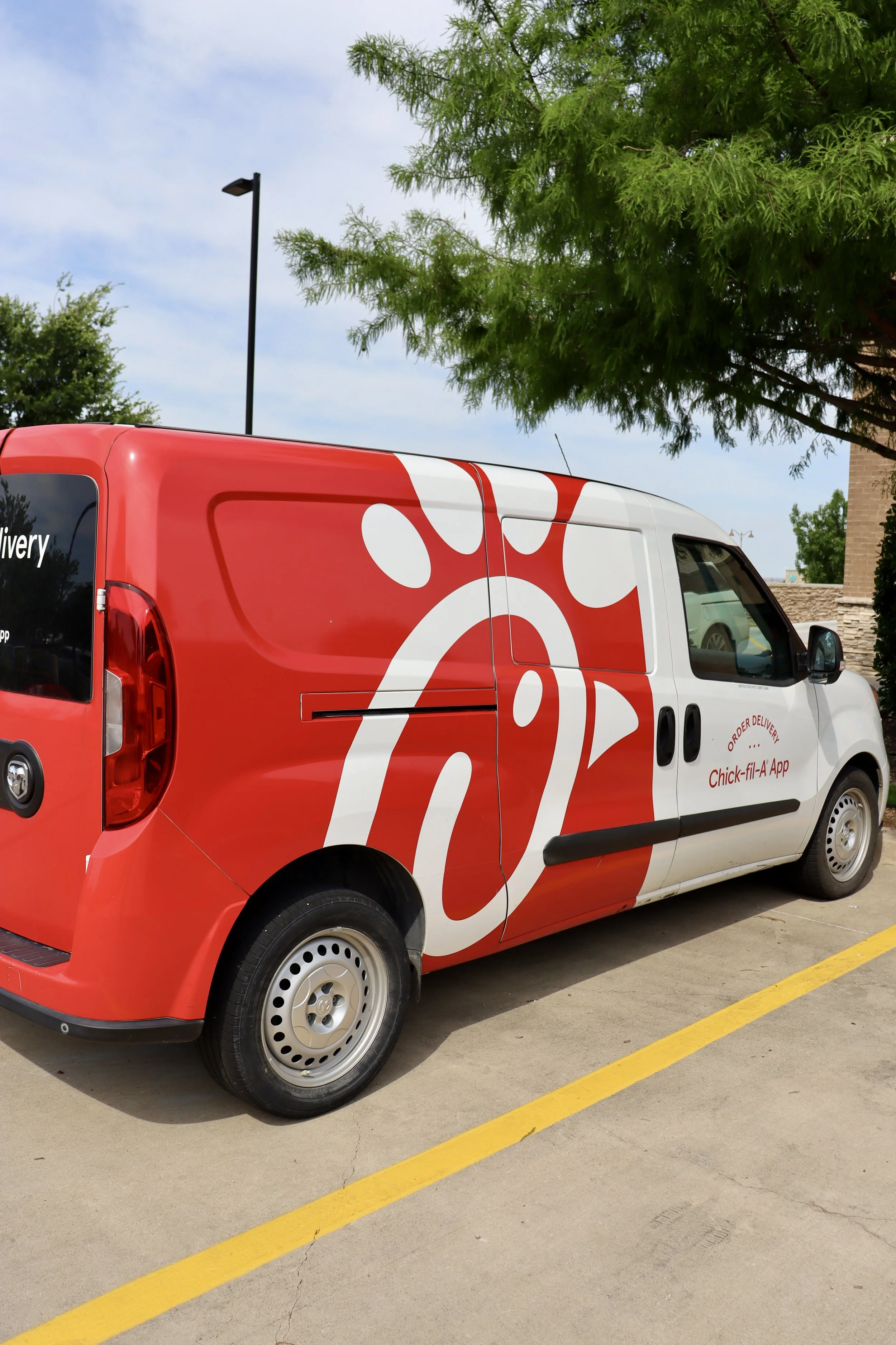 A red and white Chick-fil-A delivery van parked in a parking lot next to a green leafy tree, with a street lamp and building in the background.