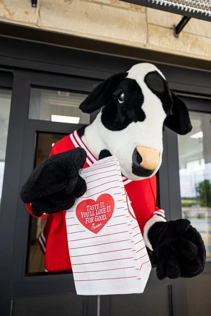 A person in a cow mascot costume is holding a Chick-fil-A paper bag with the slogan 'Taste it. You'll love it for good.'