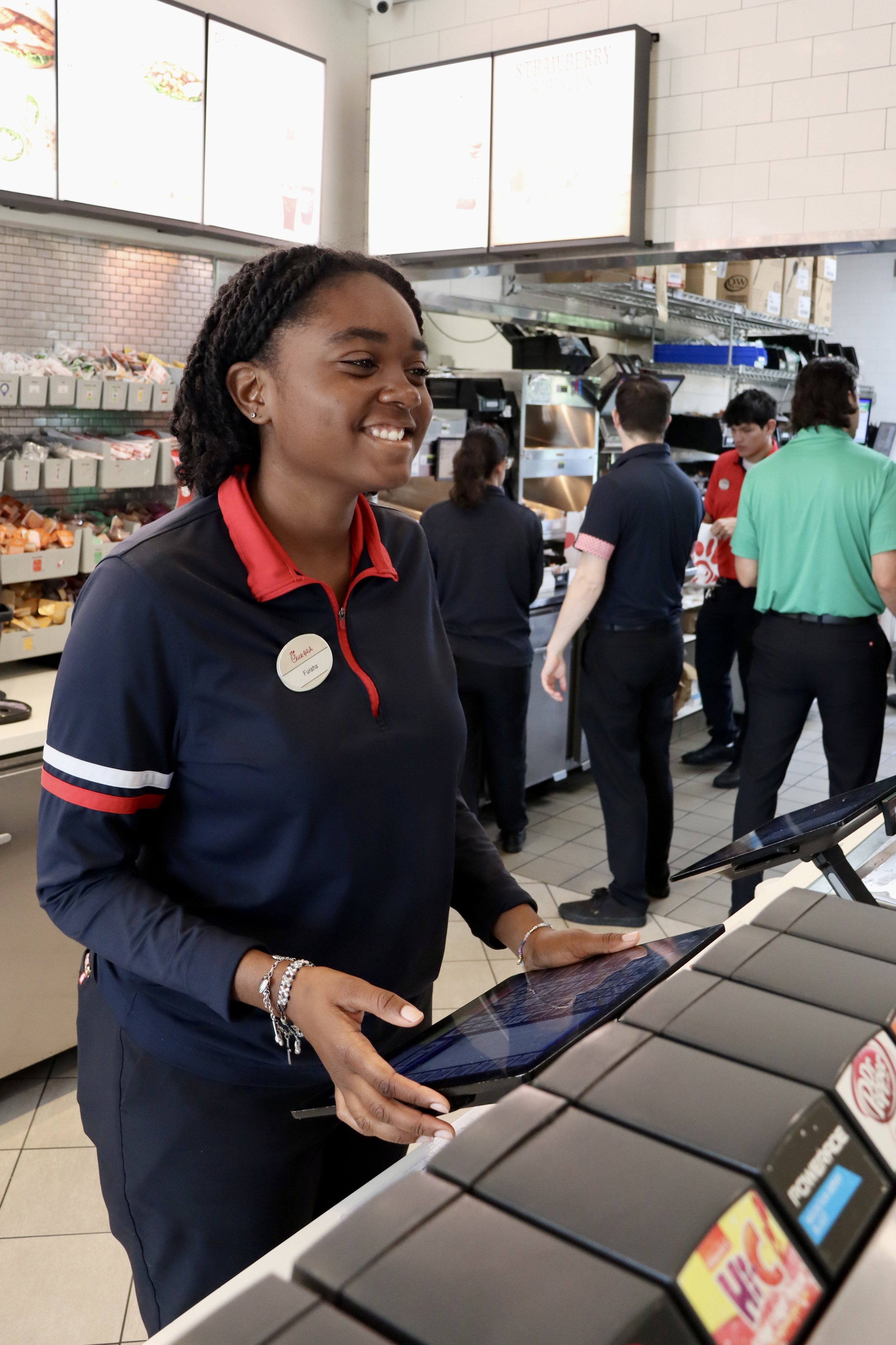 A smiling female employee at a fast food restaurant register, with other employees working behind her, in a casual dining setting.