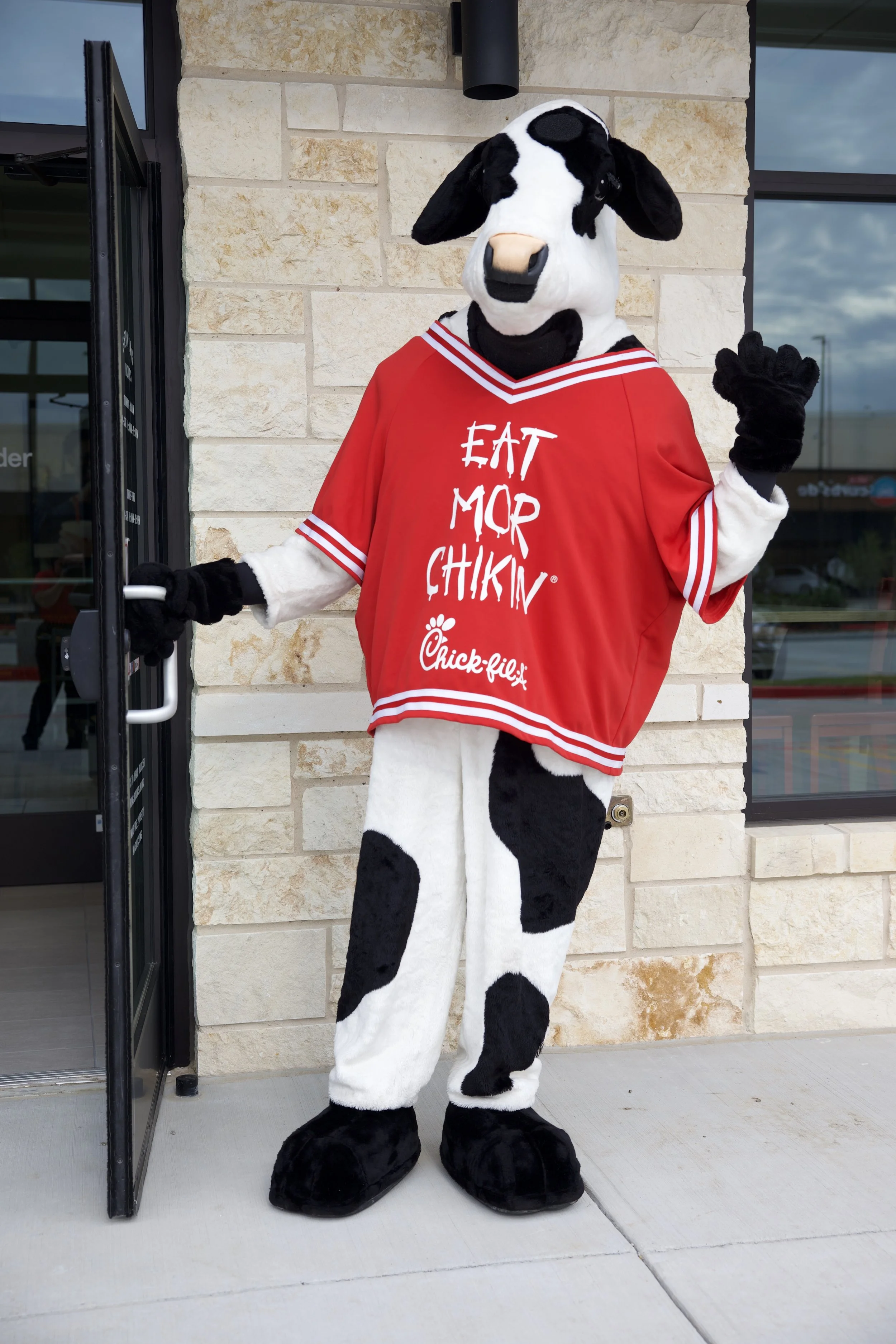 A person in a cow mascot costume wearing a red Chick-fil-A jersey standing outdoors in front of a light-colored brick wall, holding the door open with one hand and waving with the other.