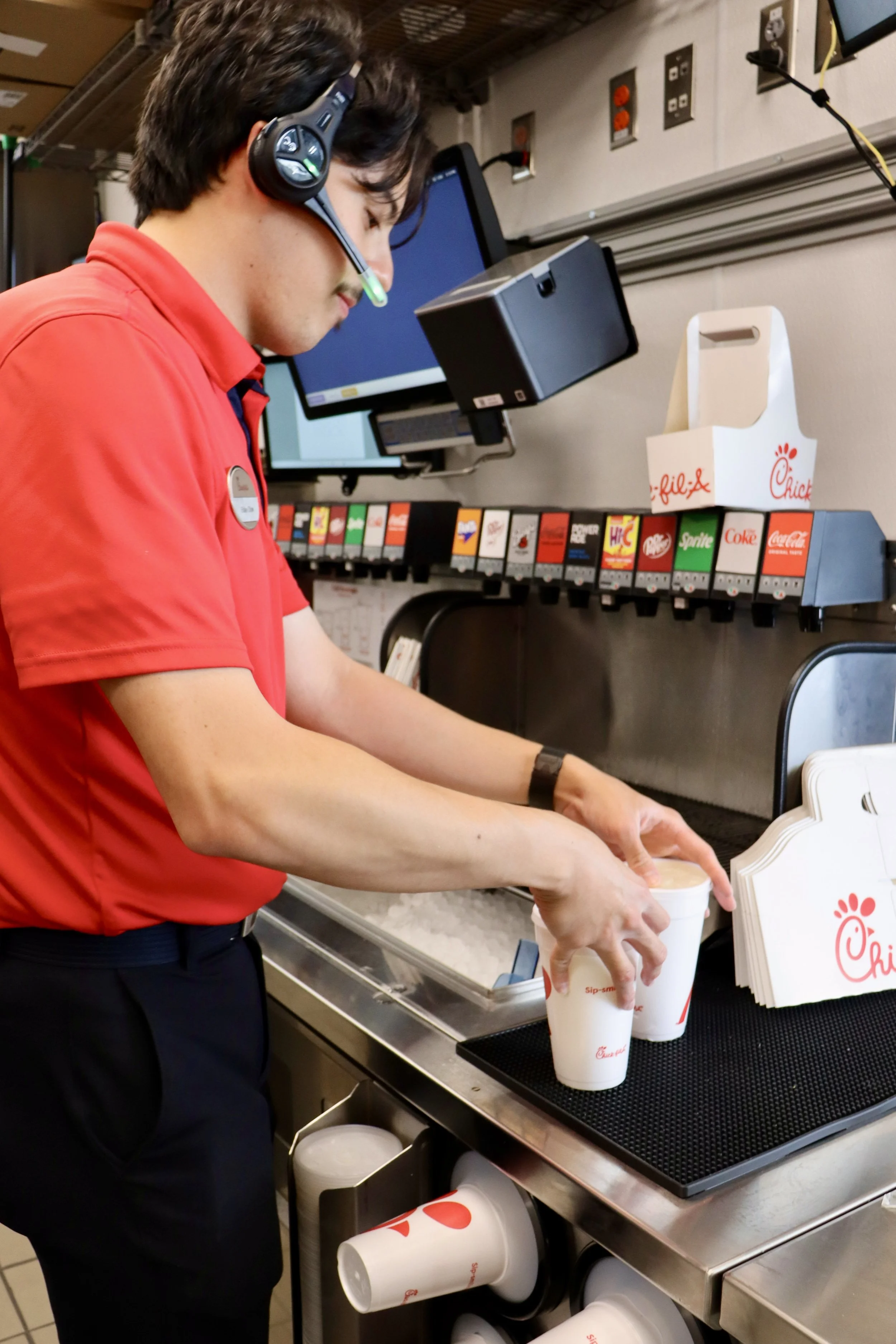 A person wearing a red shirt and black pants is filling a large Chick-fil-A branded drink cup with a beverage at a counter in a fast-food restaurant. The person is wearing a headset with a microphone.