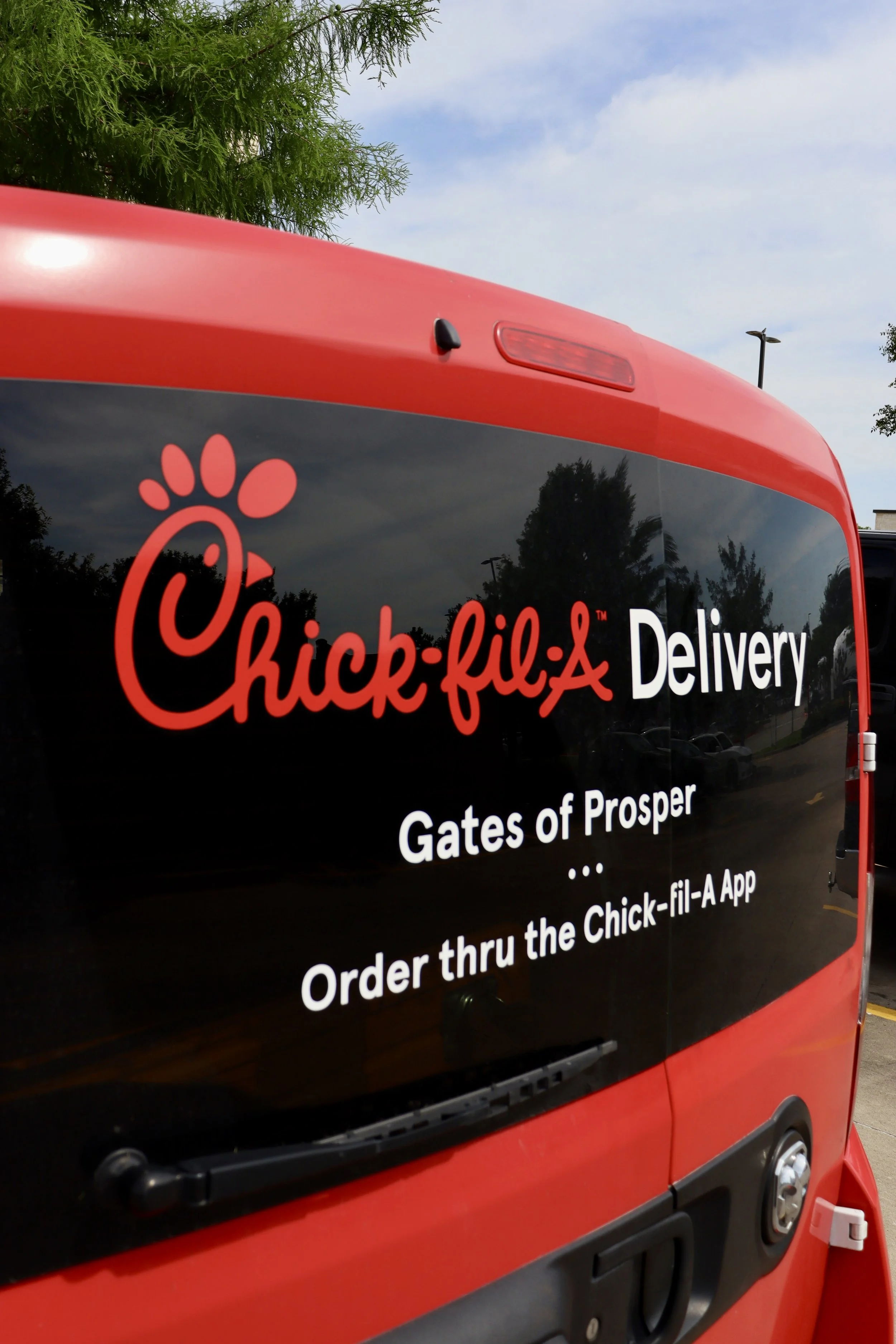 Red Chick-fil-A delivery van in a parking lot with the Chick-fil-A logo and text on it, and some trees and a lamppost in the background.