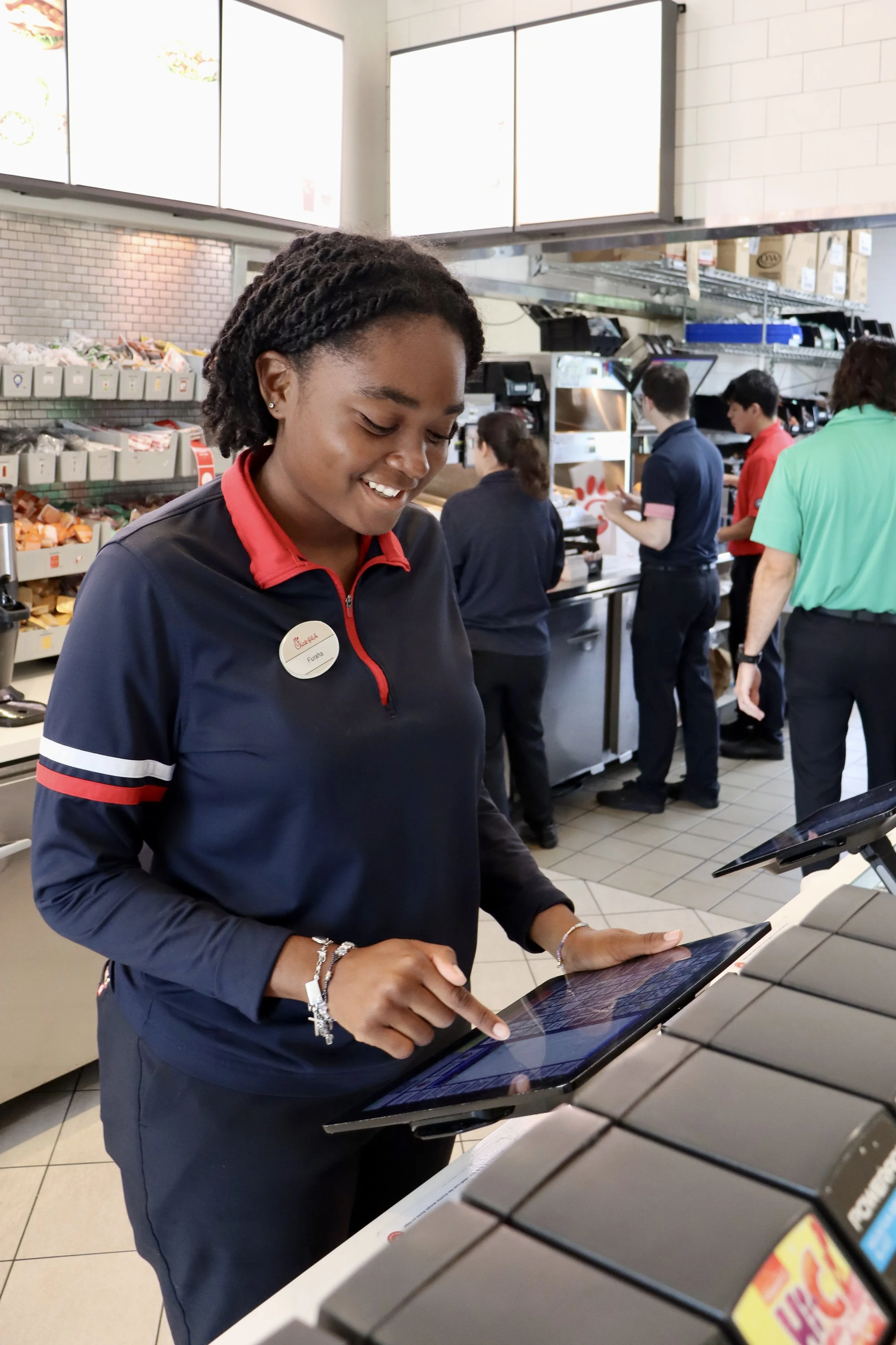 A smiling employee wearing a navy uniform with red accents and a name tag, using a touchscreen at the register in a fast-food restaurant, with other employees working behind the counter.