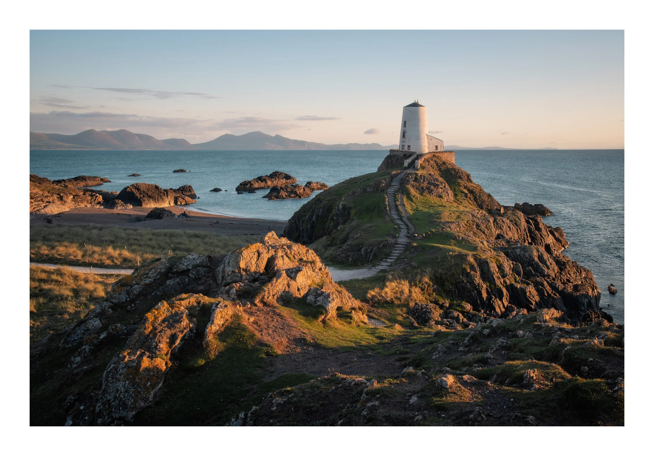 A lighthouse on a rocky hill overlooks the ocean at sunset, with a mountain range in the background.