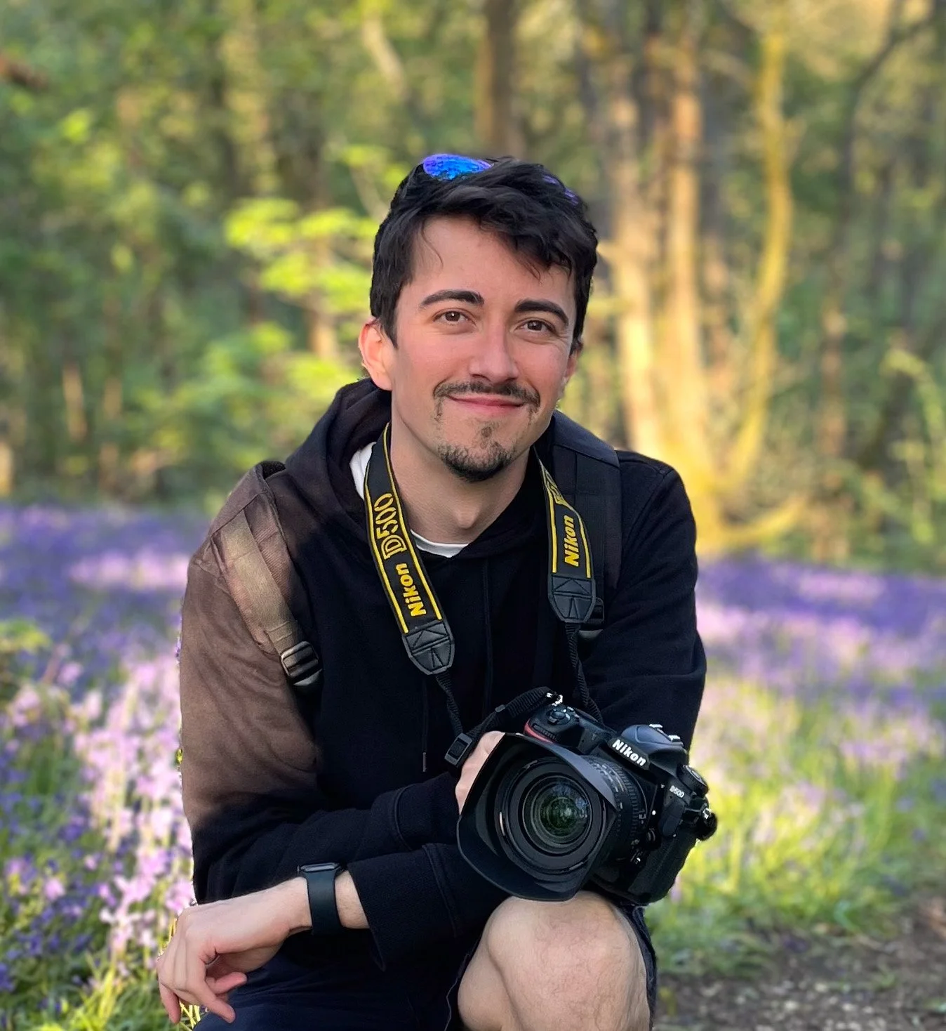 A man with dark hair and a goatee, holding a Nikon camera, sitting in a forested area with purple flowers and trees in the background.