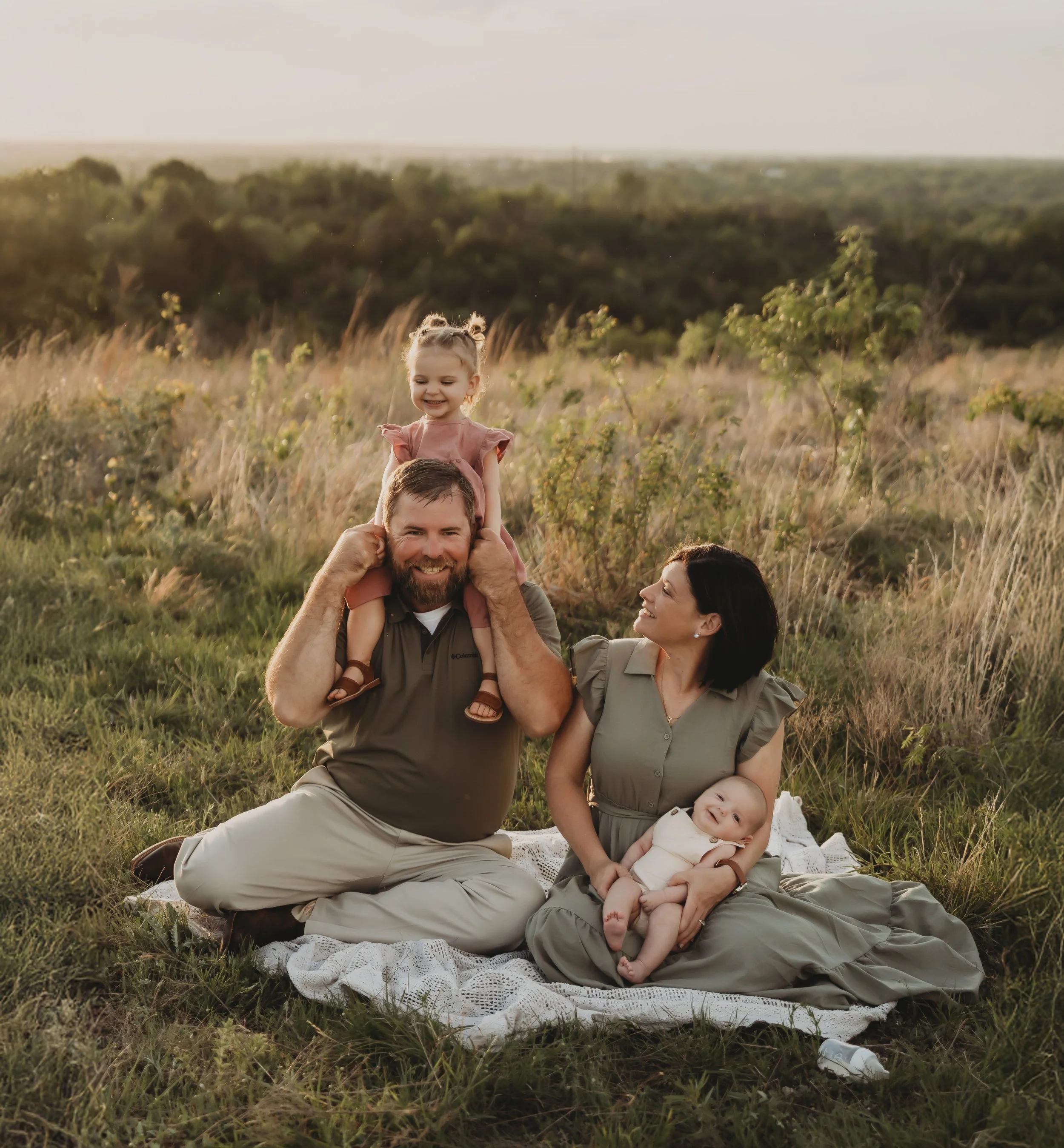A family of four enjoying a picnic outdoors in a grassy field during sunset. The father, is sitting on a blanket with their young daughter on his shoulders. The mother, is sitting on the blanket with their baby, smiling at her. The background features trees and a distant horizon.