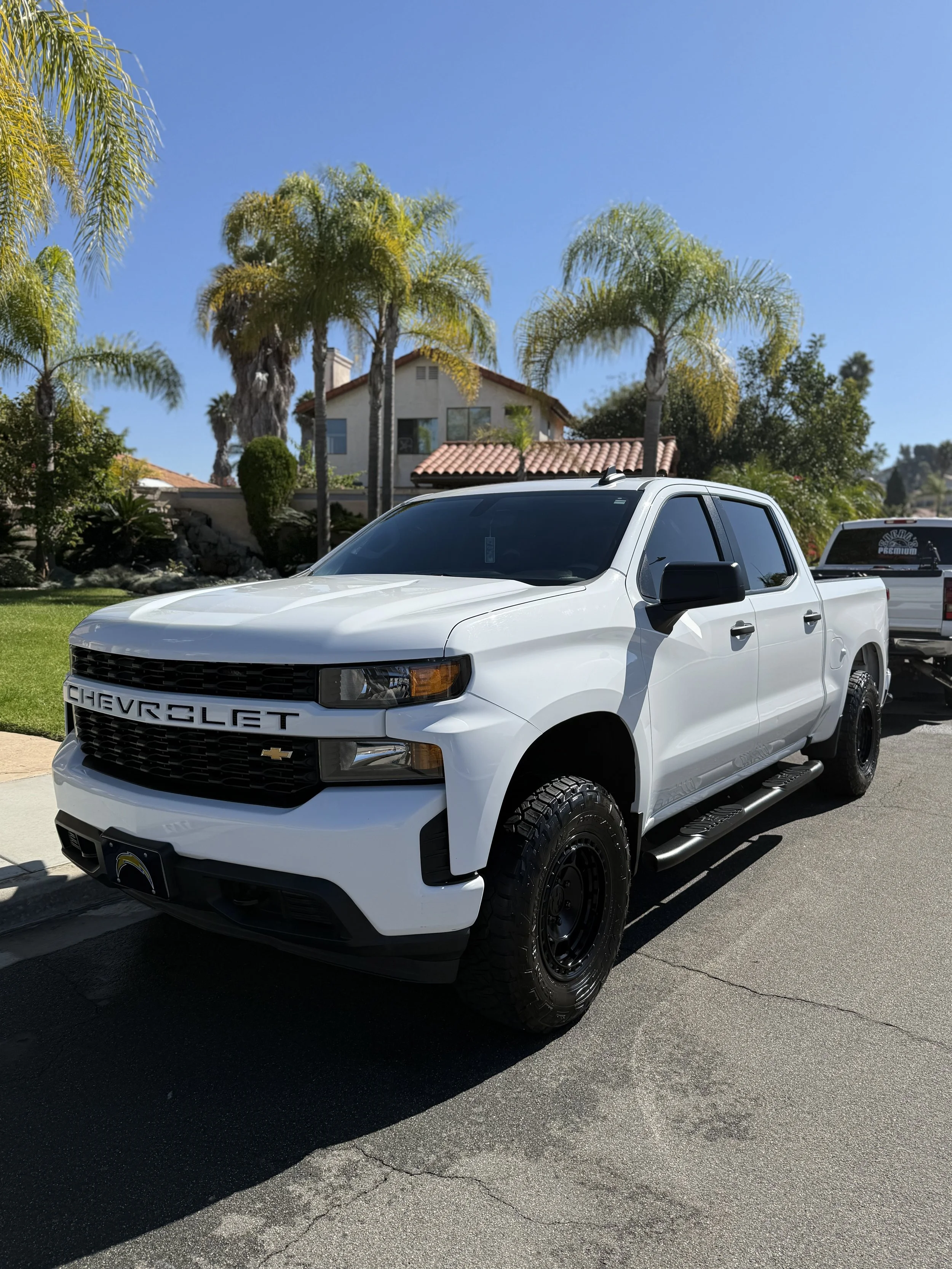 White Chevrolet pickup truck parked on a residential street with palm trees and a house in the background.