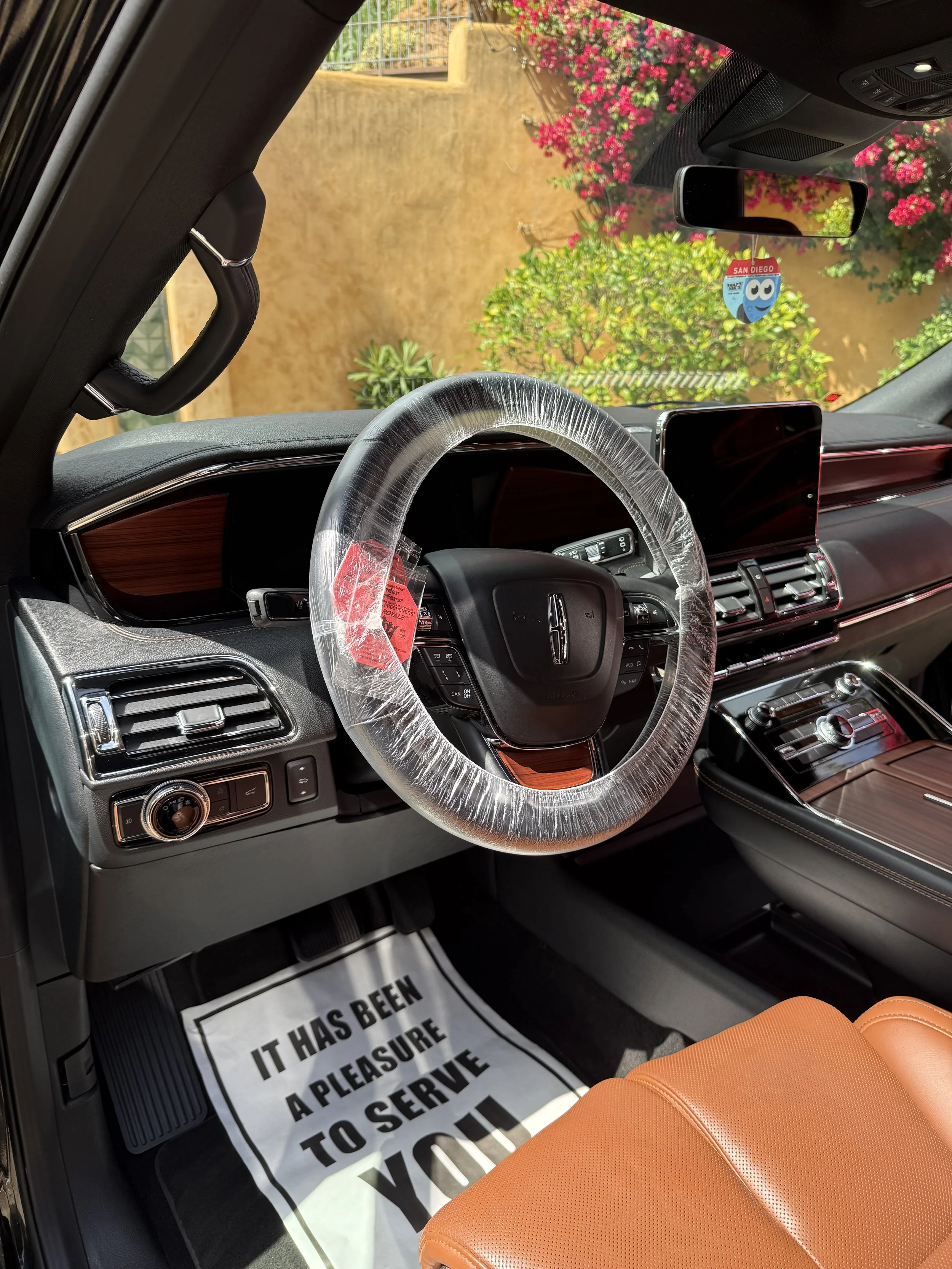 The interior of a car showing the dashboard, steering wheel with a transparent cover, orange seat, and a paper floor mat with black text. Outside the windshield, pink flowering bushes and a beige wall are visible.