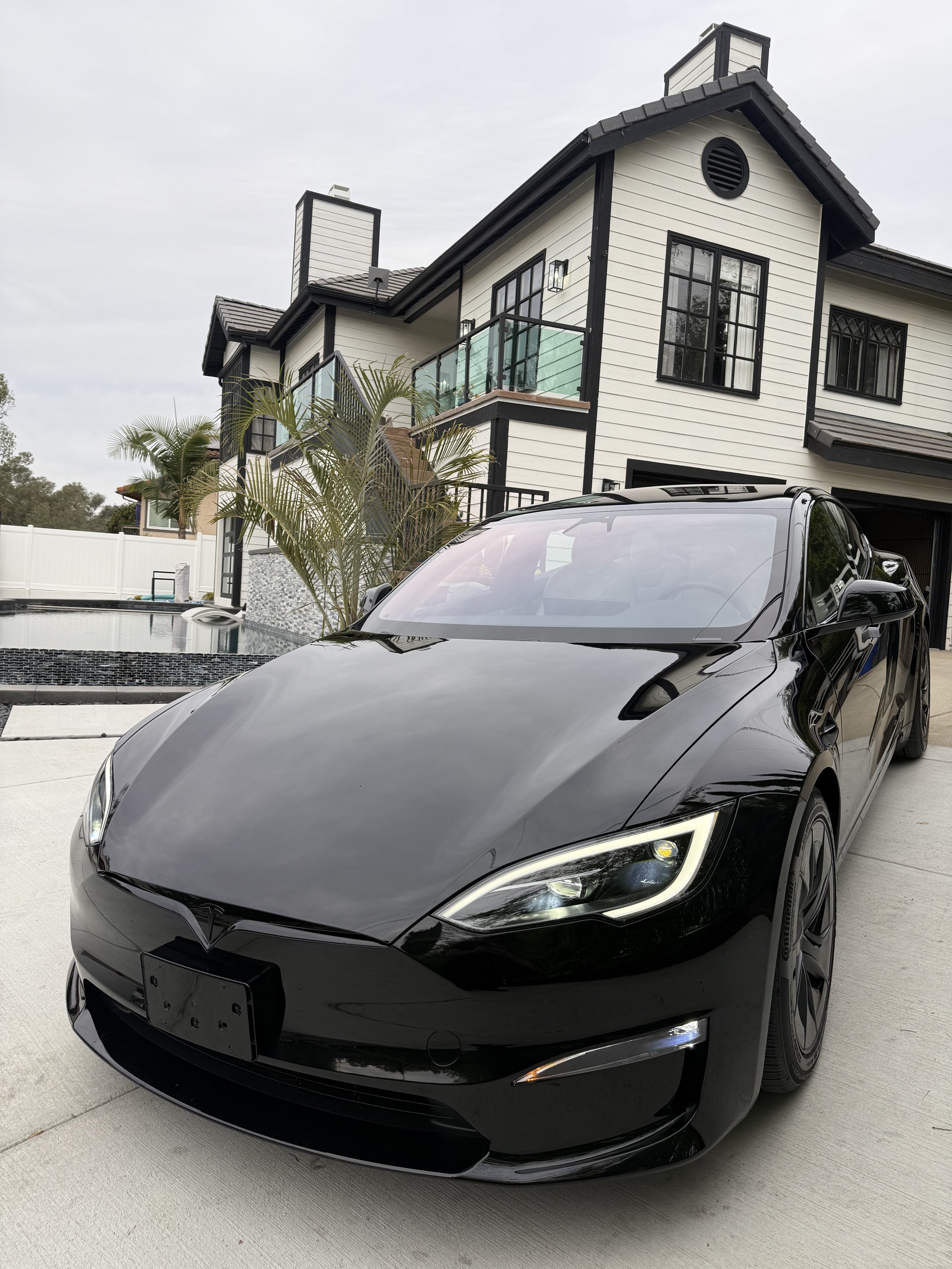 Black Tesla car parked in driveway in front of a modern house with white siding and black trim, palm plants nearby.