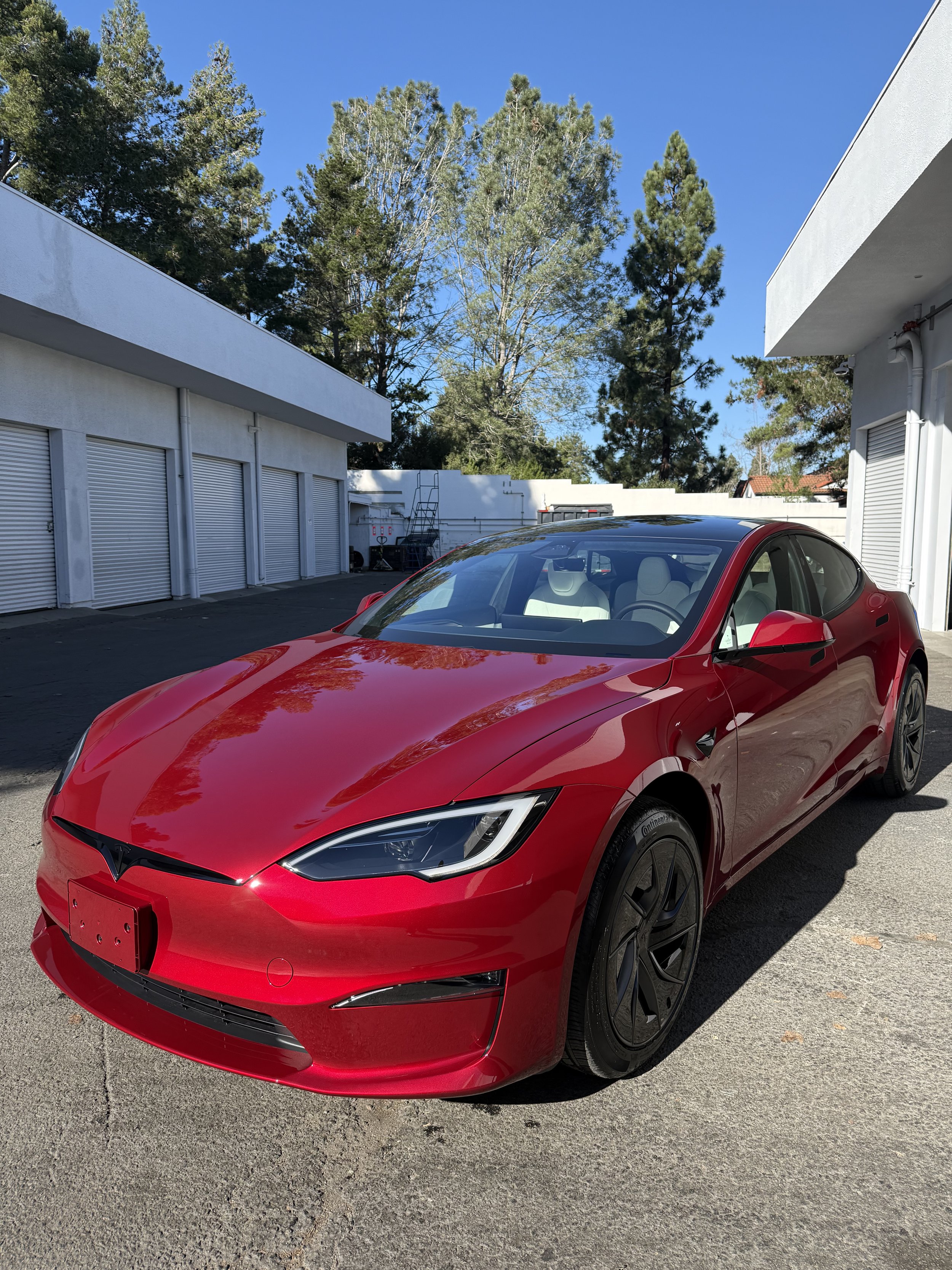 A red Tesla Model S parked outdoors on a sunny day, with trees and white buildings in the background.