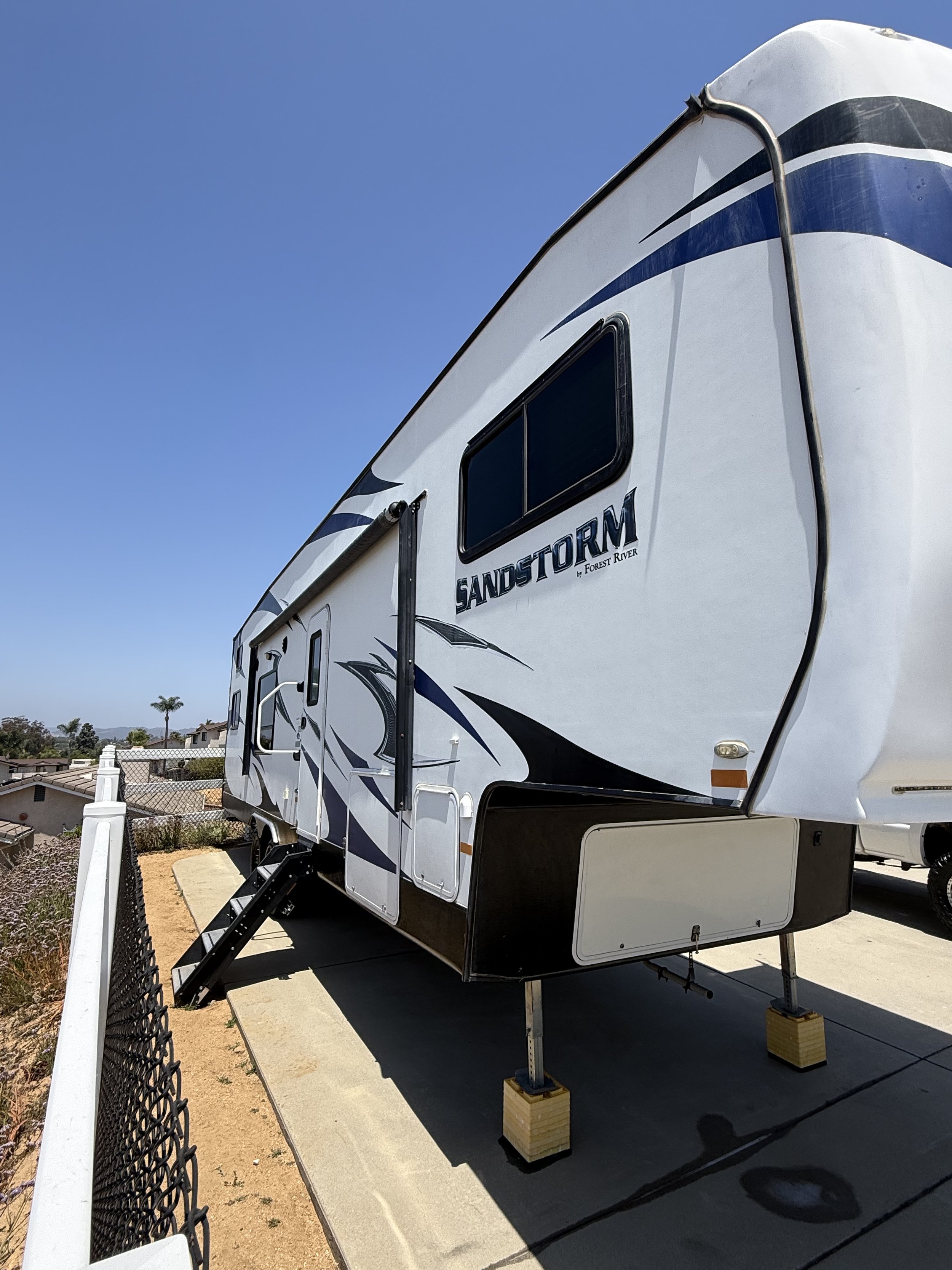 A white Sandstorm RV with black and blue graphics, parked on a concrete pad with blocks under the front jacks, next to a black chain-link fence and residential neighborhood under a clear blue sky.