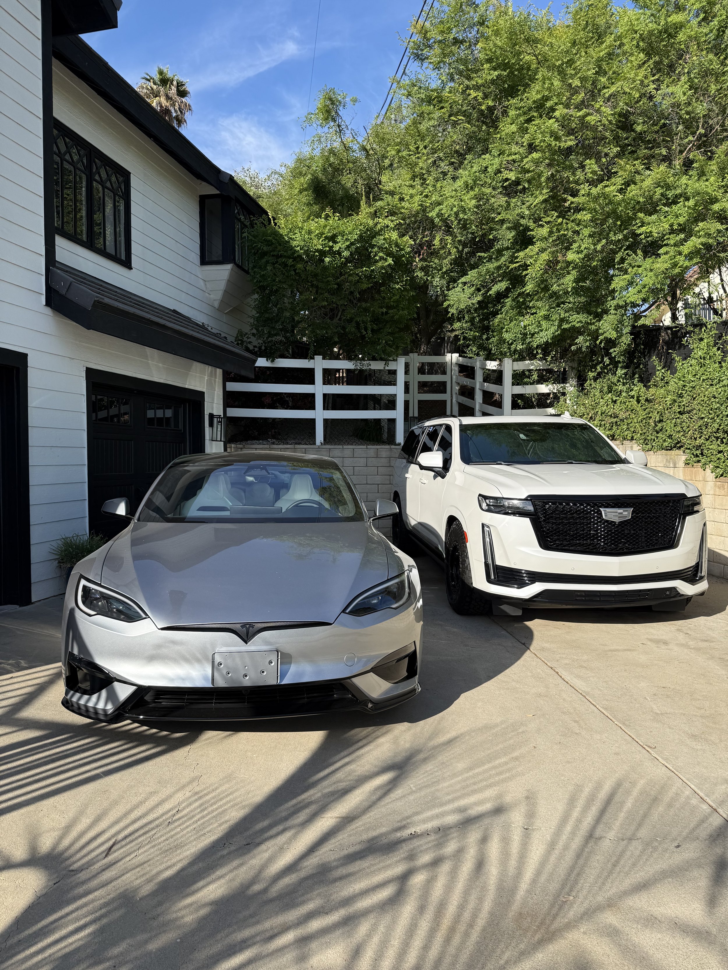 A driveway with two parked cars, one silver Tesla and one white Cadillac SUV, in front of a white house with black windows and a surrounded by green trees under a bright blue sky.