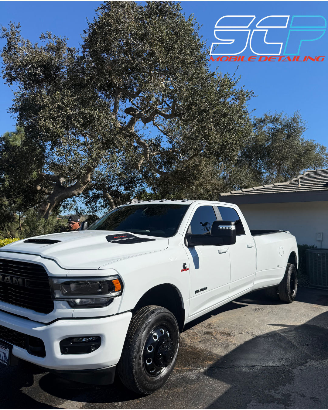 A white Ram truck parked in front of a tree and house under a clear blue sky, with a person wearing a black cap standing nearby.