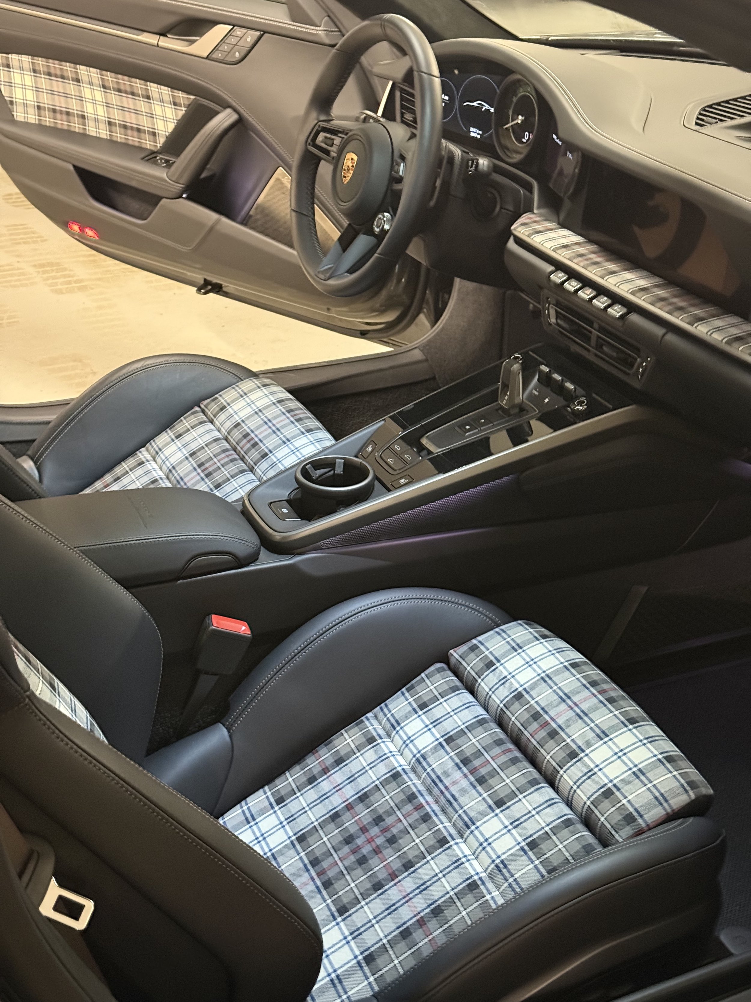 Interior of a luxury sports car featuring plaid-patterned seat upholstery, a black leather steering wheel with a Porsche emblem, and a dashboard with digital displays.