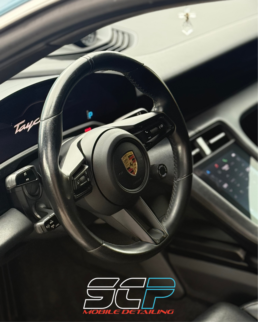 Close-up of a Porsche Taycan interior, showing the steering wheel with the Porsche logo and a digital dashboard display.