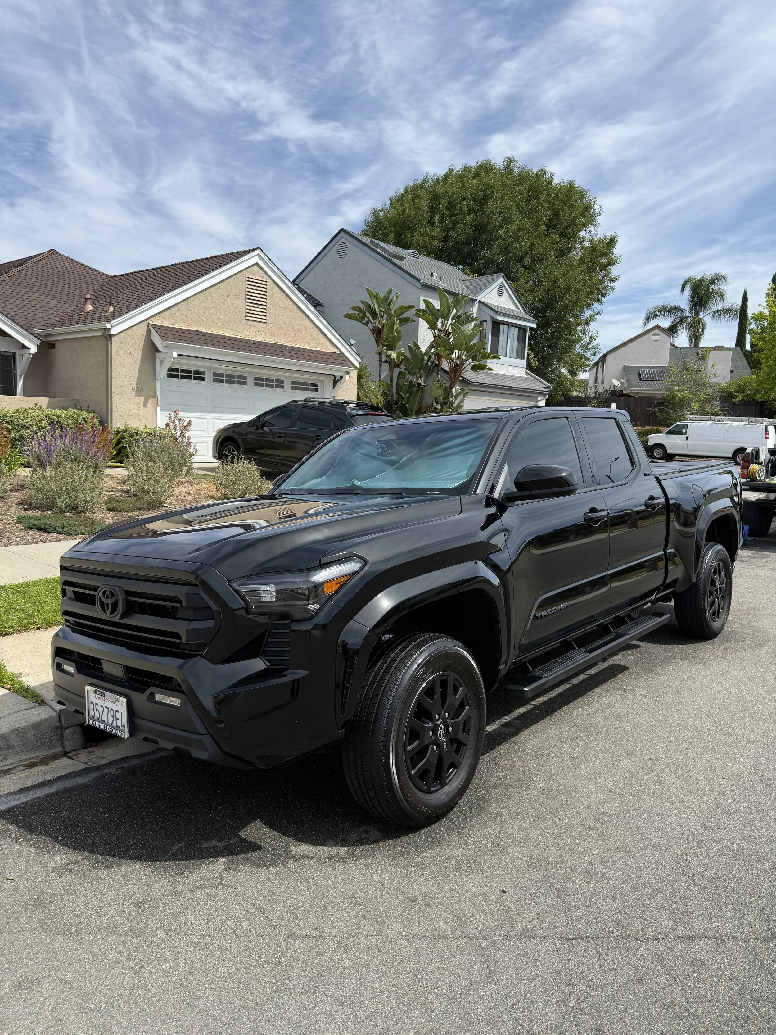 A black Toyota Tacoma pickup truck parked on a residential street in front of houses with green lawns, trees, and a partly cloudy sky.