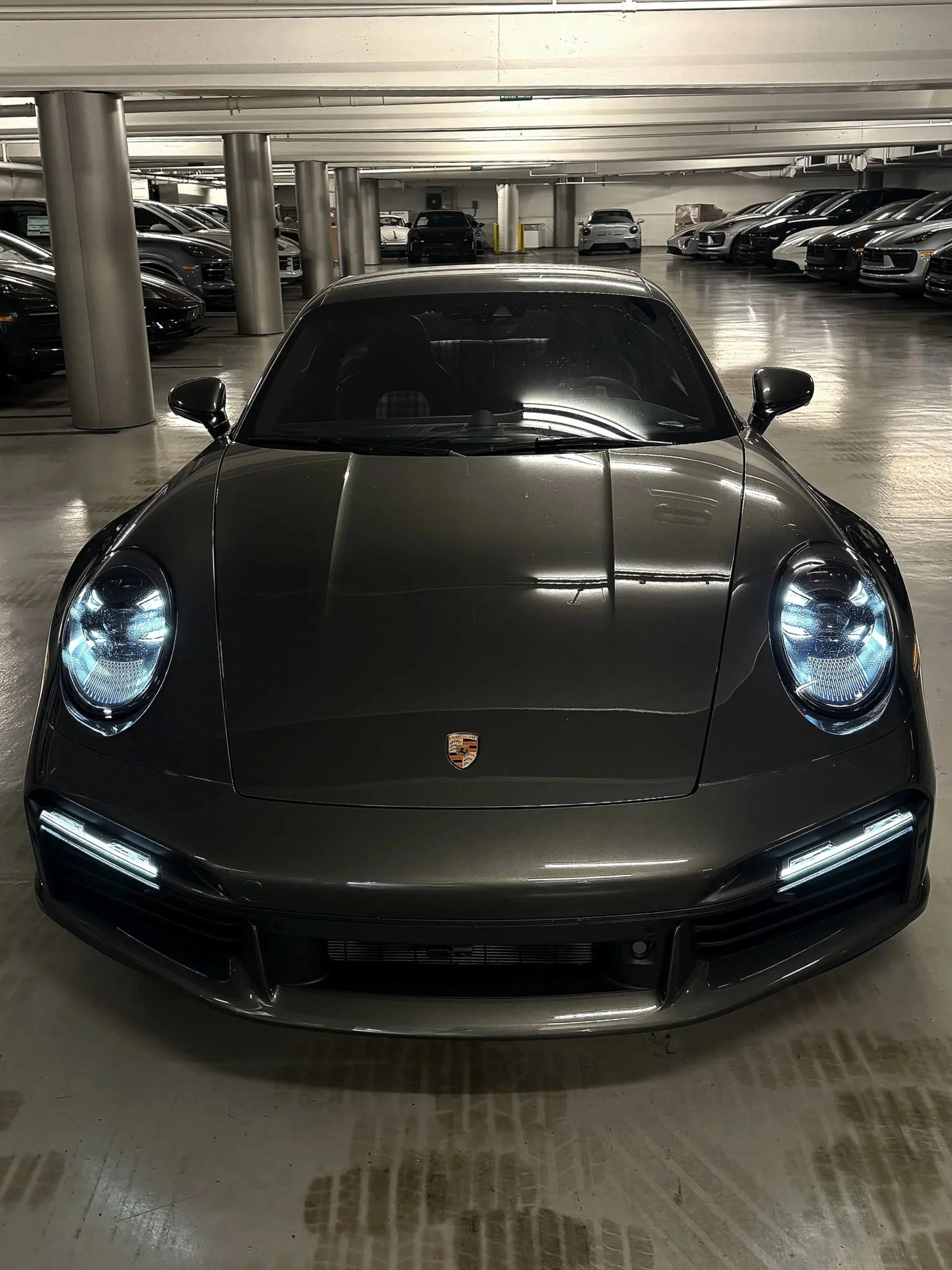 A black Porsche sports car parked in an indoor parking garage, facing forward. The garage has multiple rows of luxury cars and concrete pillars supporting the ceiling.