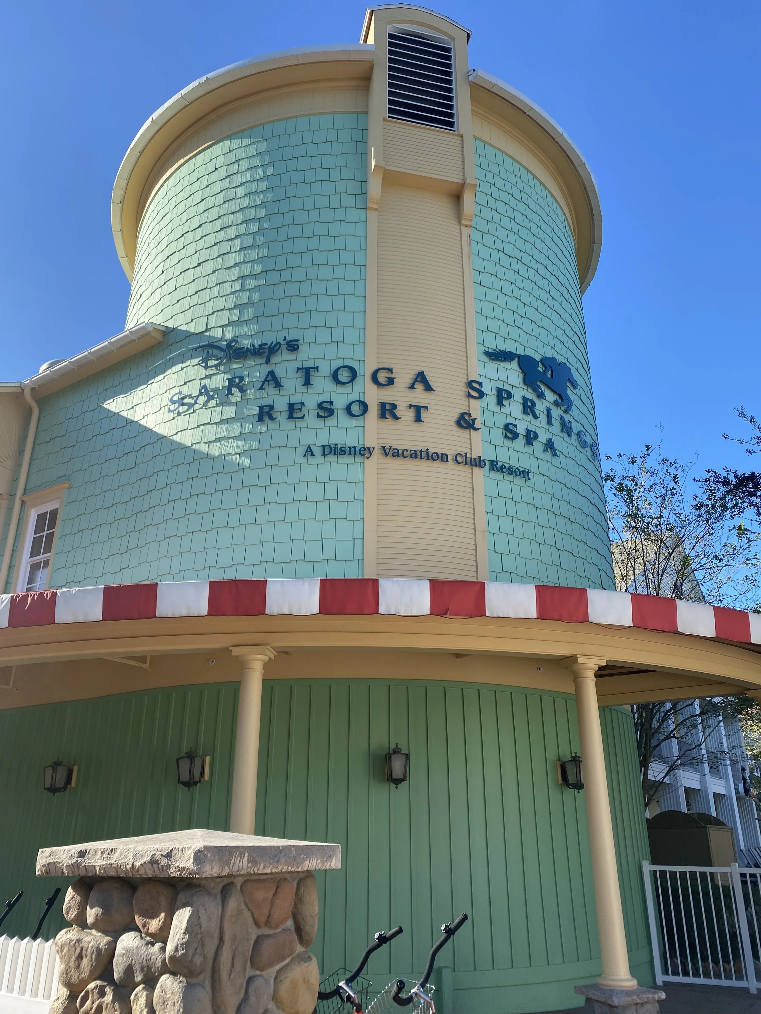 A turquoise-colored building with a sign that reads 'Disney's Saratoga Springs Resort & Spa' and features a silhouette of a horse. The building has a rounded tower with a vent at the top. In front, there is a stone well and two bicycles.