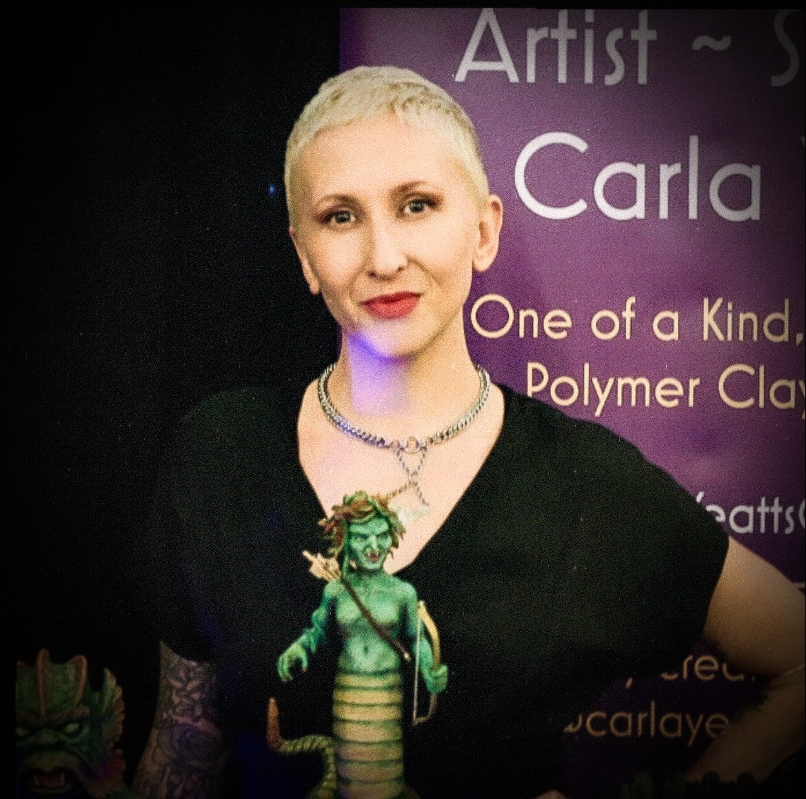Portrait of a woman with short blonde hair, wearing a necklace, black top, and a badge with a green mythical creature on it, standing in front of a purple backdrop with text about an artist named Carla and polymer clay art.