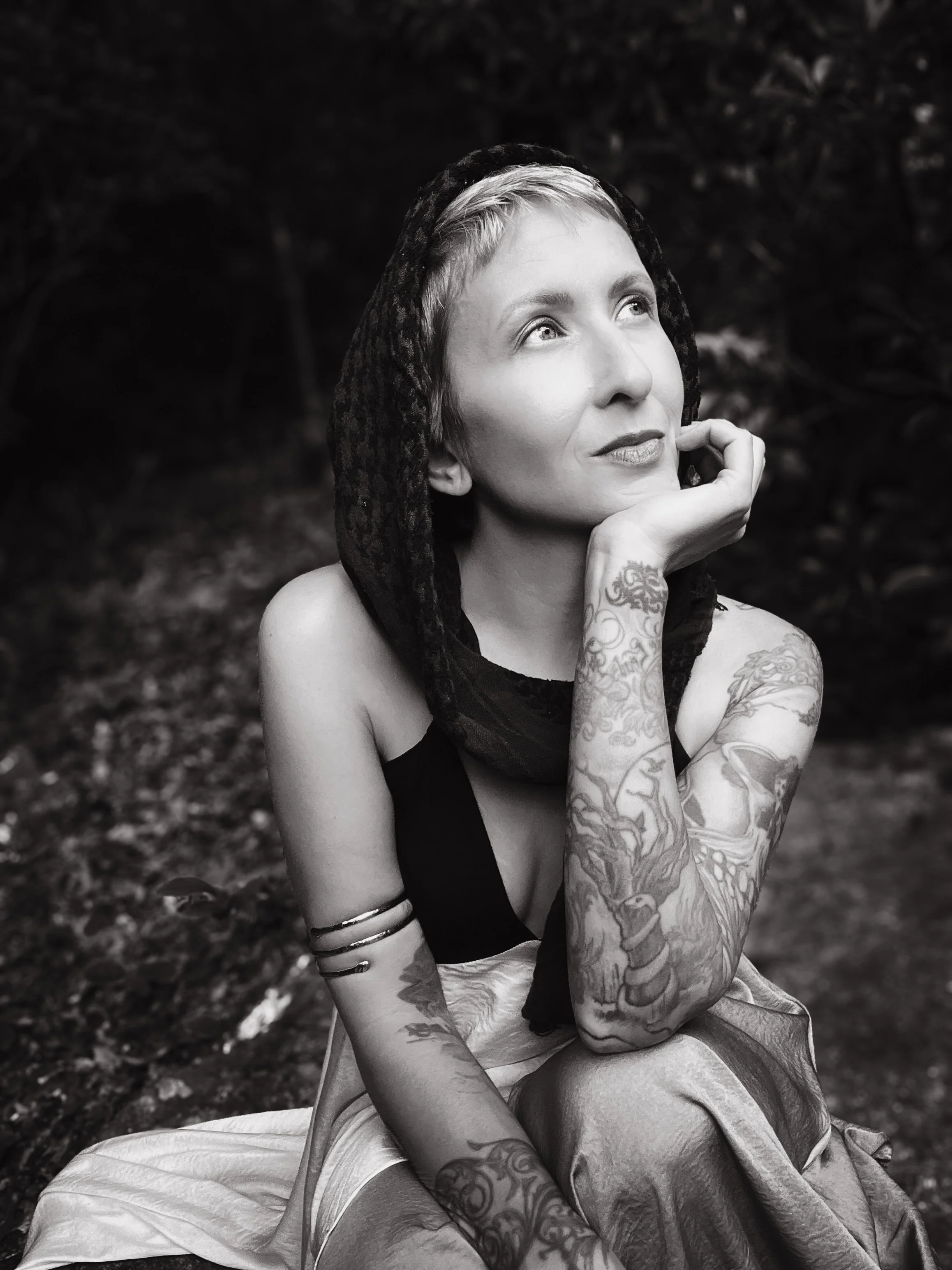 Black and white photo of a woman with short hair, wearing a black lace hood, and tattoos on her arms, sitting outdoors with a thoughtful expression, resting her chin on her hand, surrounded by trees and foliage.