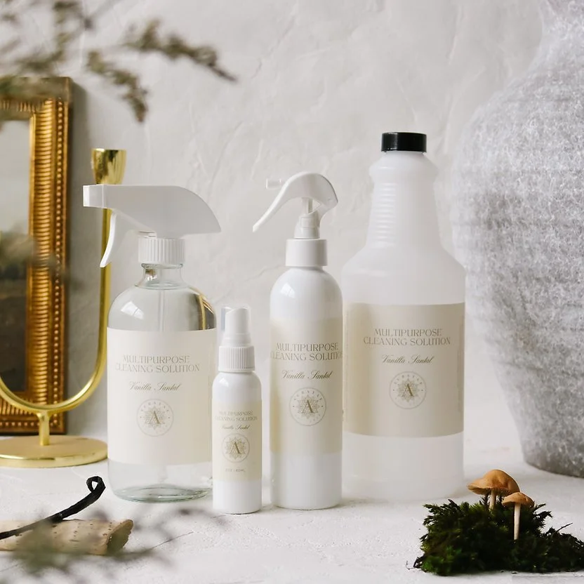 Set of cleaning spray bottles and containers labeled 'Multipurpose Cleaning Solution' with vanilla sandal scent, arranged on a white surface with a large gray textured vase, moss and small mushrooms, and a gold mirror in the background.