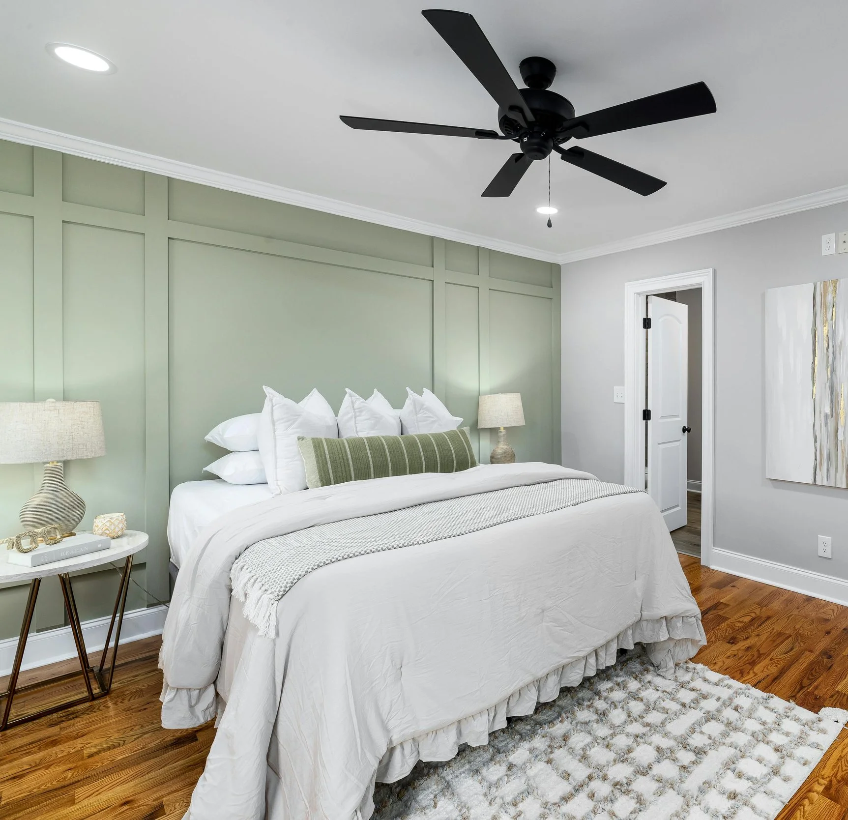 A bedroom with a bed covered in white bedding and pillows, green accent wall behind the bed, two bedside lamps, a black ceiling fan, wooden floor, a white door, and an abstract wall art.