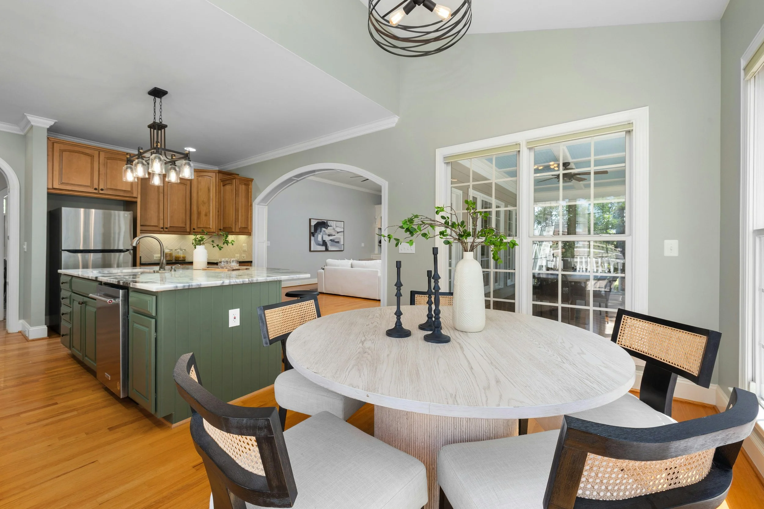 Open-concept living room and kitchen with hardwood floors, large window, round dining table with vase and candle holders, kitchen island, wooden cabinets, stainless steel refrigerator, modern light fixture, and a view into a screened porch.