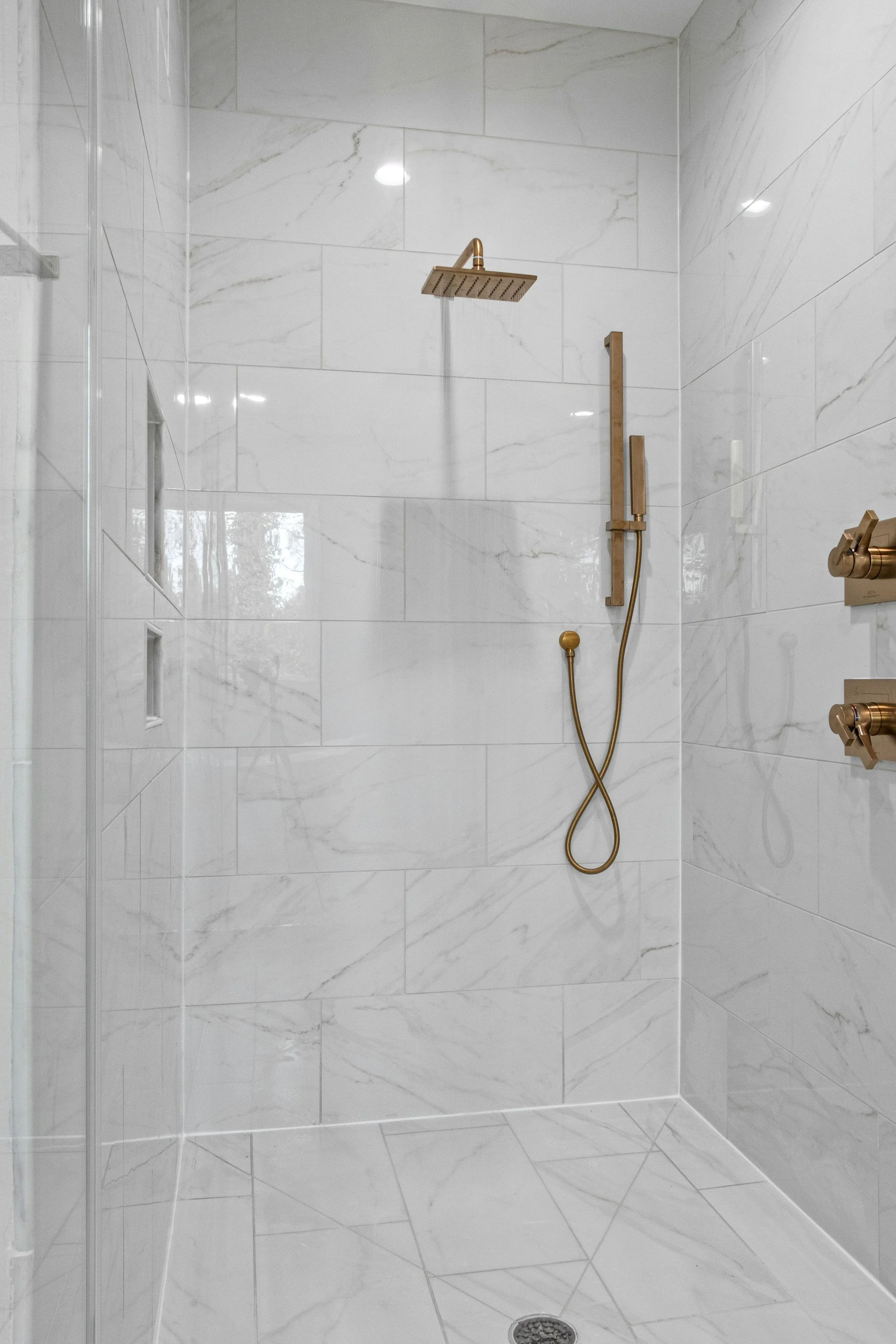 A modern shower with white marble tiles, a large square rain shower head, a handheld shower, and built-in niche shelves.
