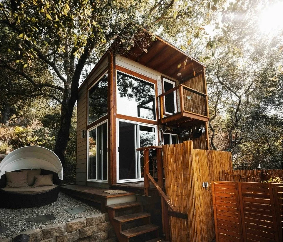 A modern two-story tiny house with large windows and a small upper balcony, surrounded by trees, with an outdoor seating area featuring a cushioned circular lounge. Wooden stairs lead up to the house, which is fenced with bamboo and wood materials.