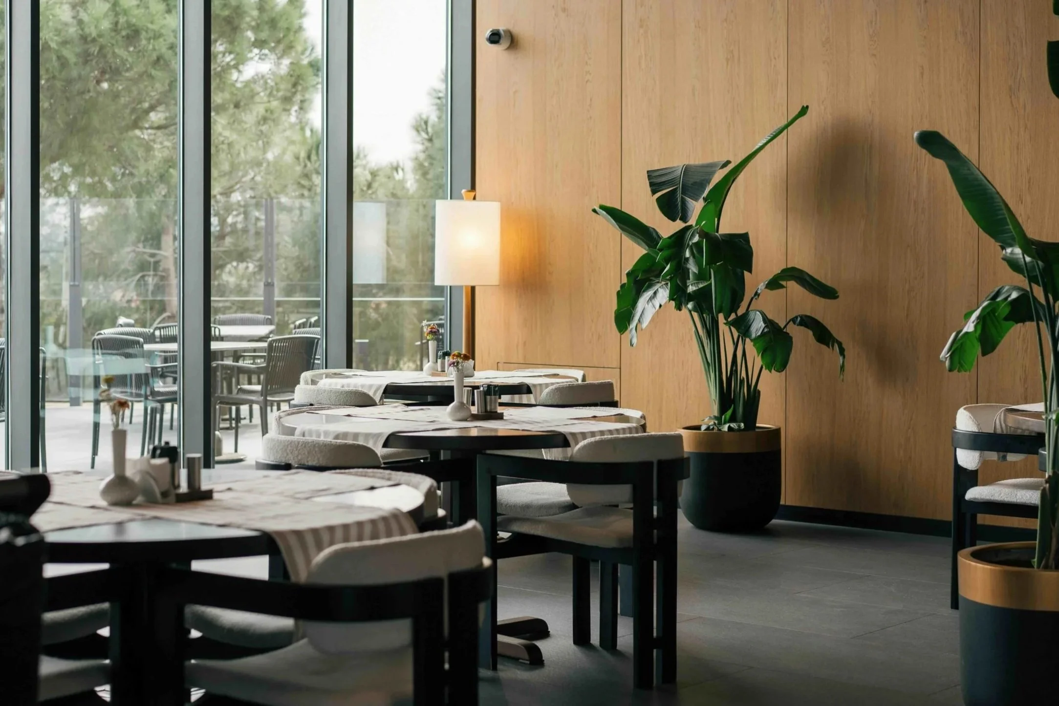 A modern dining area with multiple round tables covered with white tablecloths, decorated with small flower arrangements, and gray chairs around them. Large floor-to-ceiling windows allow natural light to fill the room, indicating an outdoor patio with chairs and tables. The interior features a wooden wall with a large green plant in a round pot, a tall white lamp, and a black security camera mounted on the wall.
