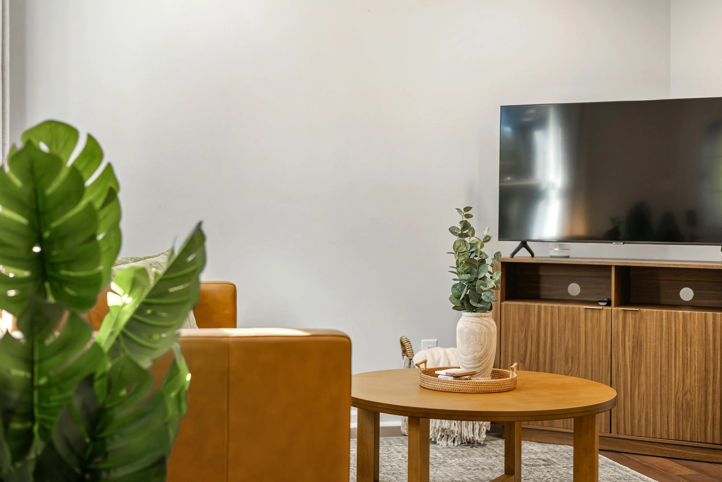 Living room interior with a large flat-screen TV on a wooden stand, a round wooden table with a woven tray and a decorative vase, green plants including a large monstera leaf and a smaller leafy potted plant, a beige armchair, and a white wall in the background.
