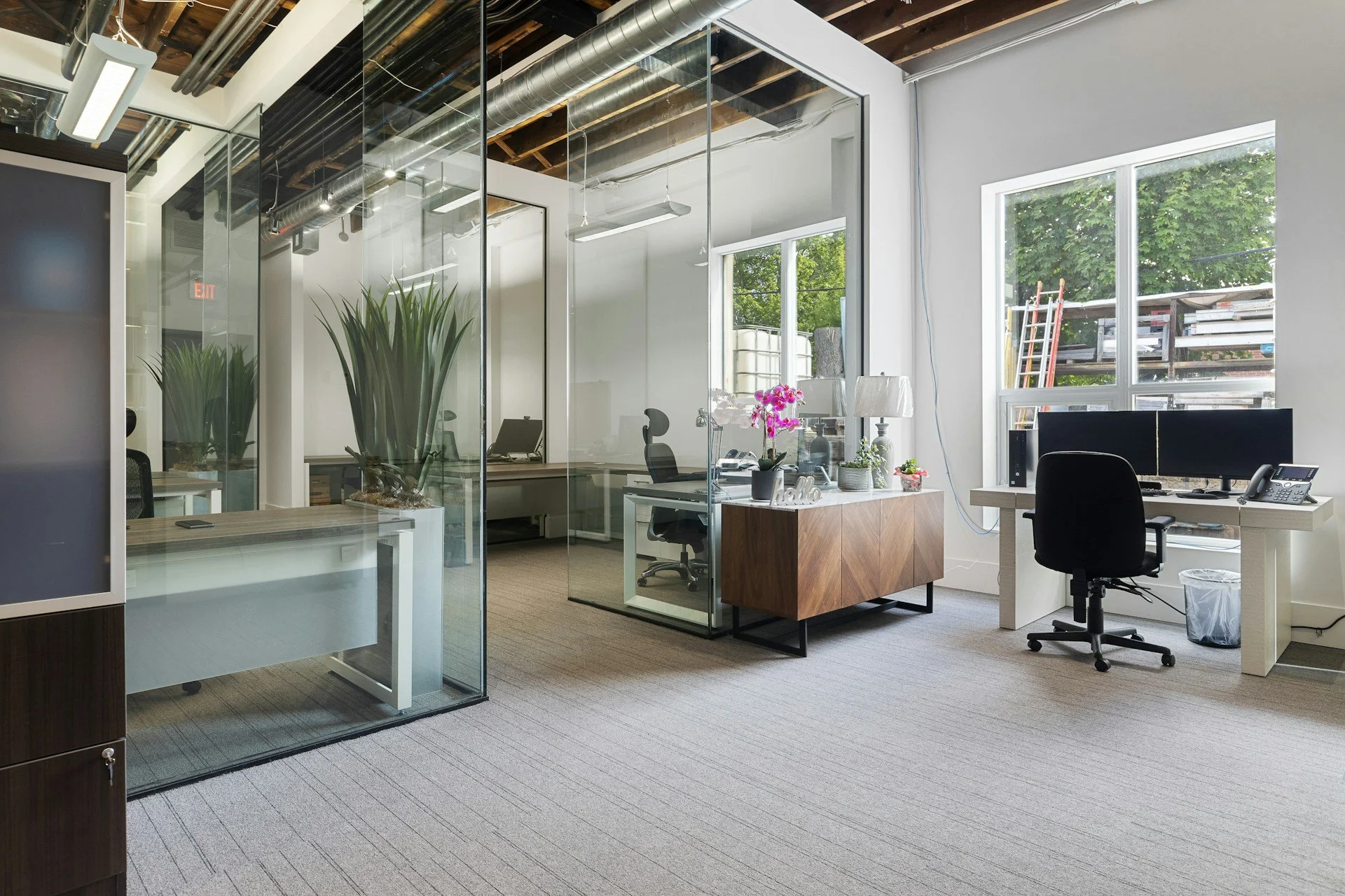 Modern office space with glass-enclosed meeting room, plants, desks with computer setups, large windows, and daylight coming into the room.