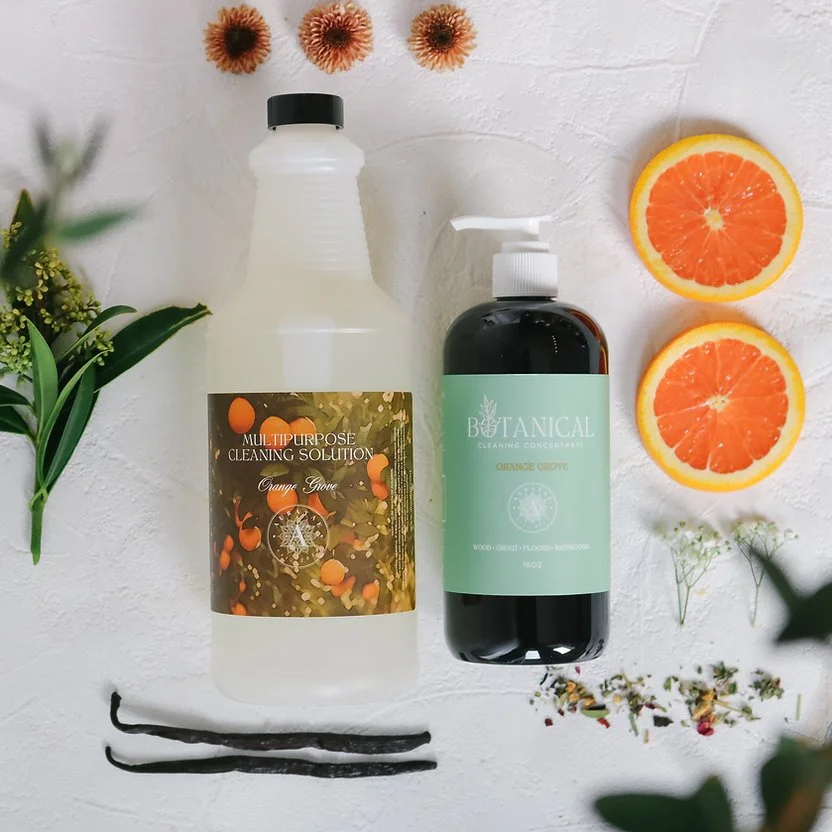 Cleaning supplies including a multipurpose cleaning solution bottle labeled 'Orange Grove' and a botanical cleaning concentrate labeled 'Orange Grove,' with slices of orange, vanilla pods, and various flowers around them on a white surface.
