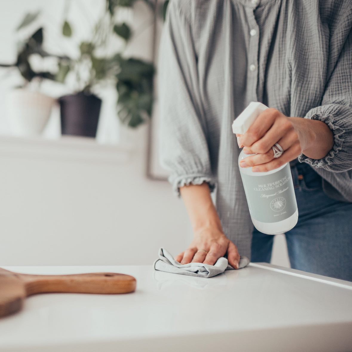Person cleaning a white countertop with a spray bottle of multi-purpose cleaning solution and a cloth, wearing a gray checkered shirt.