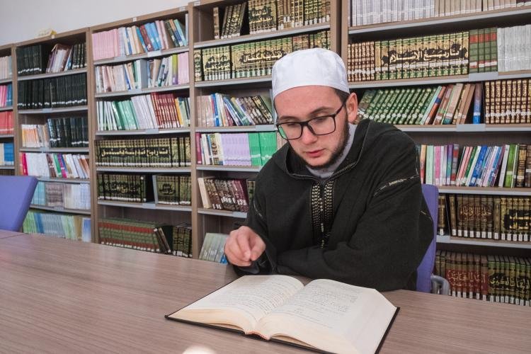 A young man wearing glasses, a white cap, and a black jacket reading a book at a wooden table in a library with shelves of books behind him.