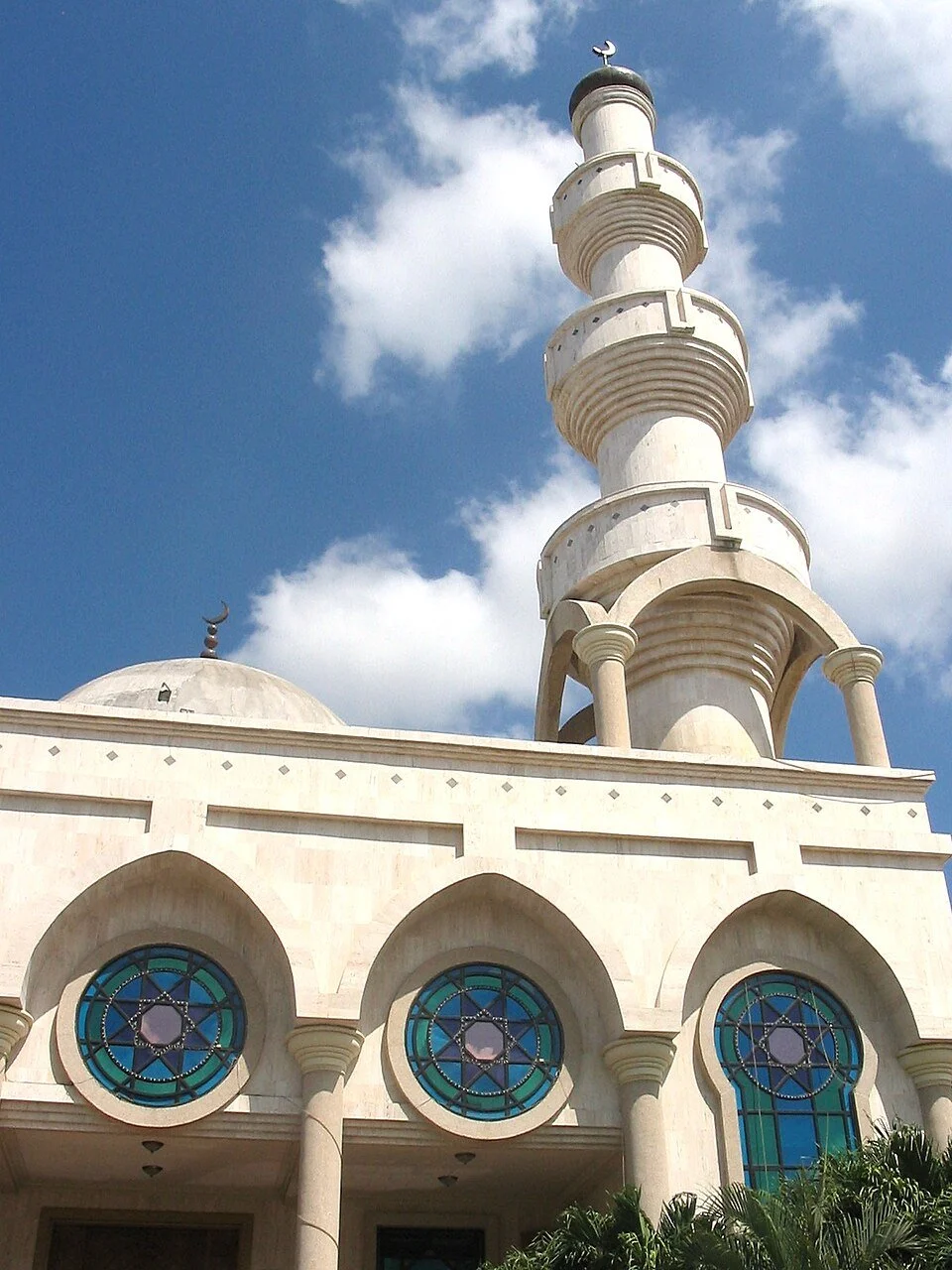 A mosque with a tall minaret and blue stained glass windows, under a partly cloudy sky.