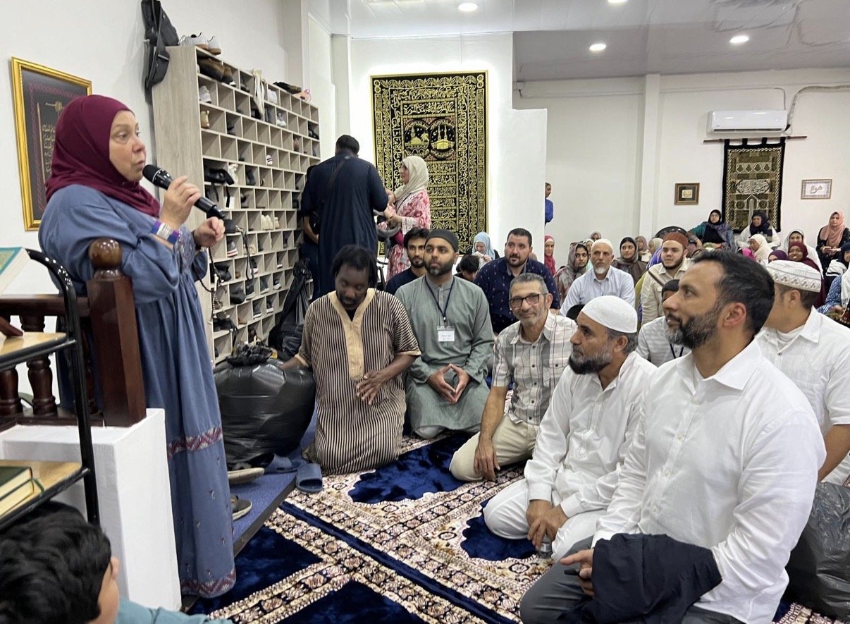 A woman in a maroon headscarf and long blue dress speaking into a microphone while others listen, seated on a prayer rug in a room with shelves and Islamic decorations.