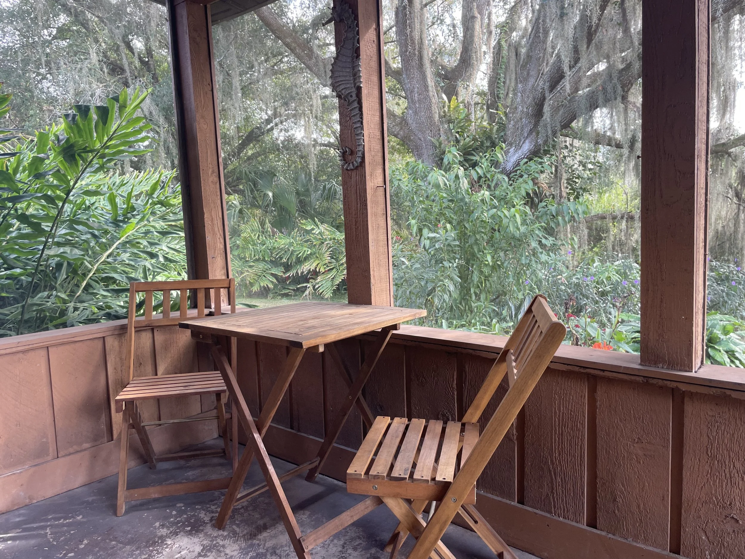 A screened porch with wooden furniture including a table and chairs, overlooking a lush, green garden with large trees and various plants.